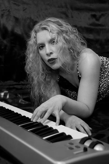 Woman with curly hair playing piano, looking at camera, in black and white, wearing patterned top.