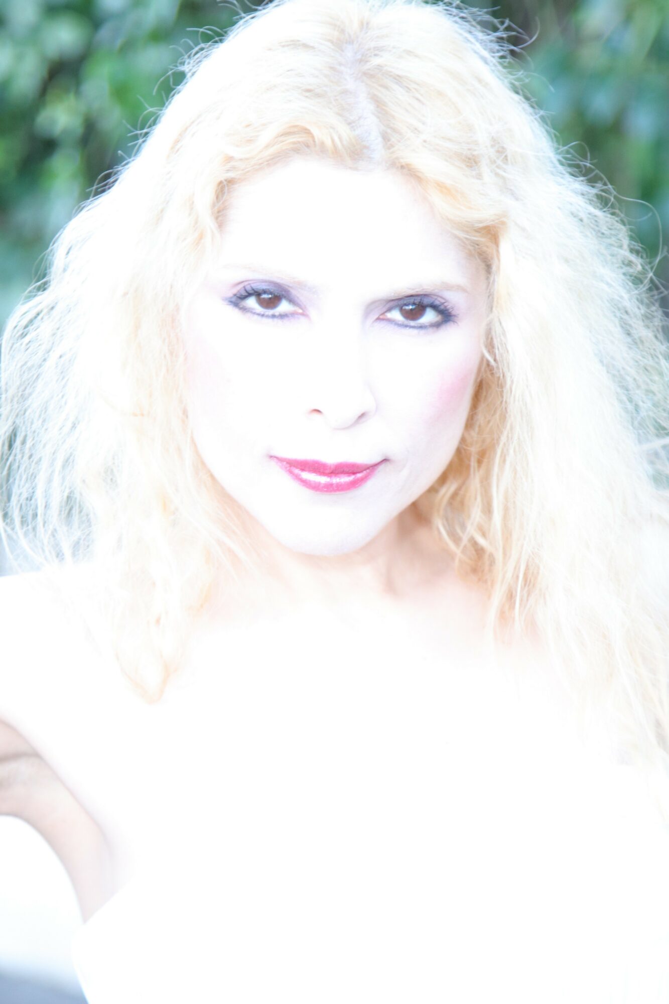 Close-up of a woman with light blonde hair and makeup, outdoors with greenery in the background.
