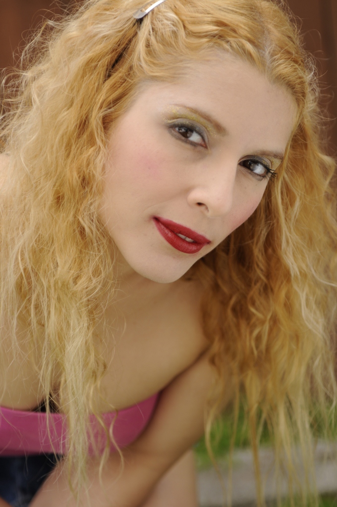 Close-up of a woman with curly blonde hair, red lipstick, and makeup, looking at the camera.