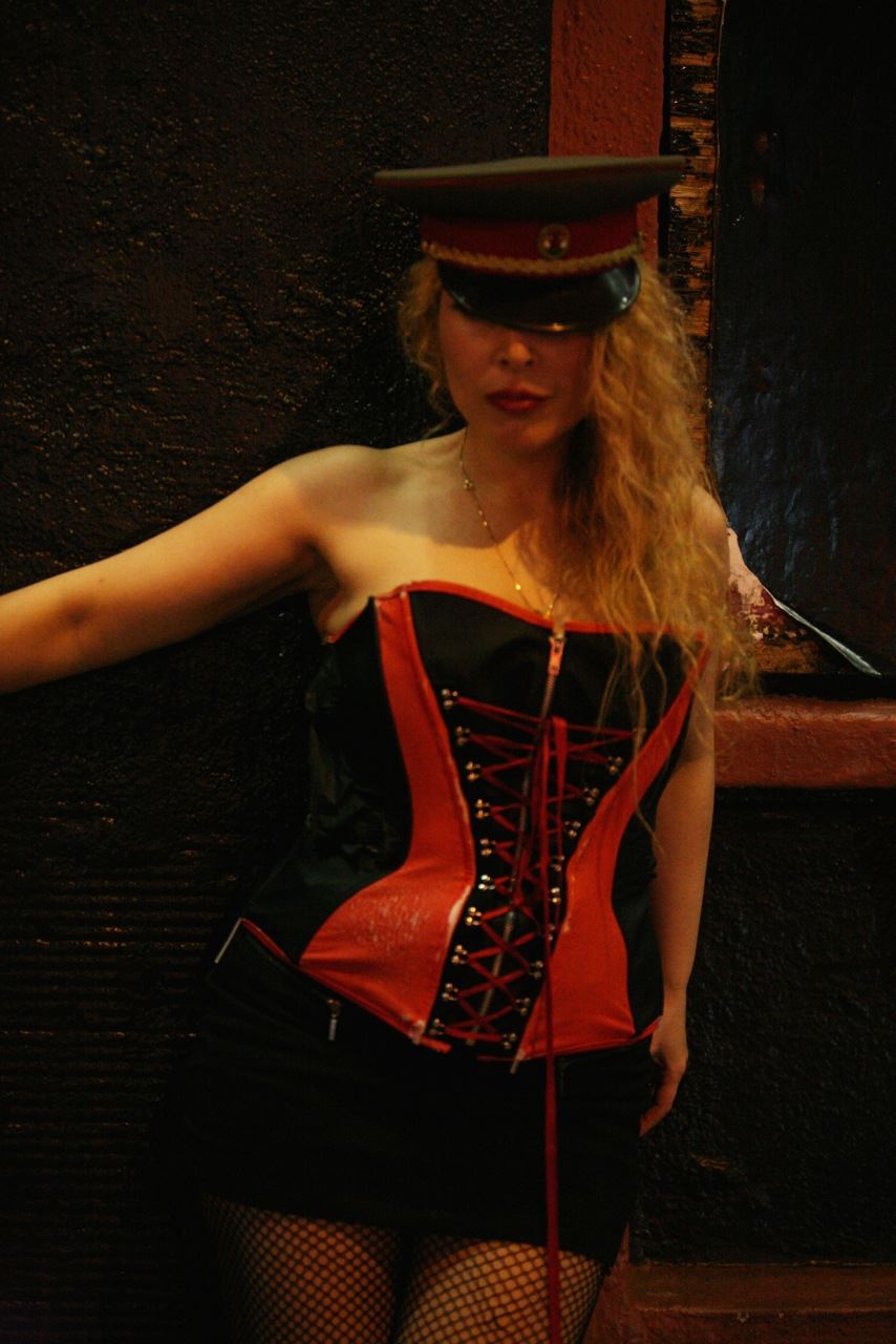Woman in a costume with a hat, corset, and fishnet stockings, standing against a dark background.