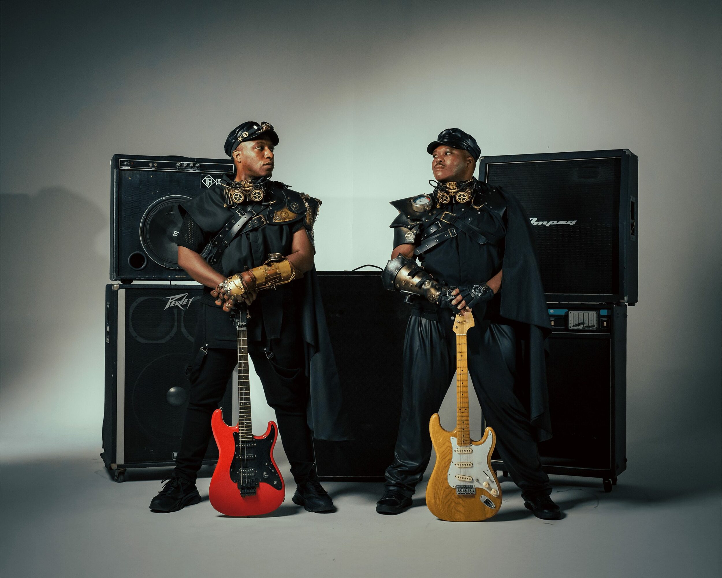 Two musicians in black outfits with gold accents stand with guitars and speakers in a studio setting.
