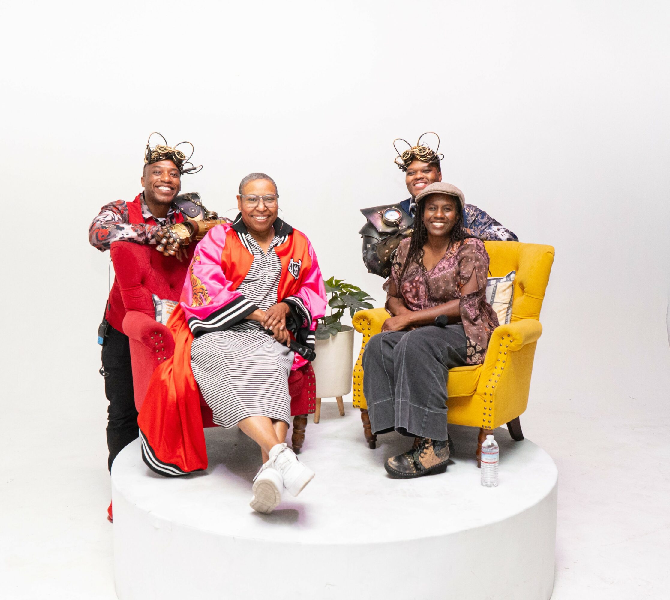 Four women sitting and standing on a white circular platform, smiling, with a plain white background.