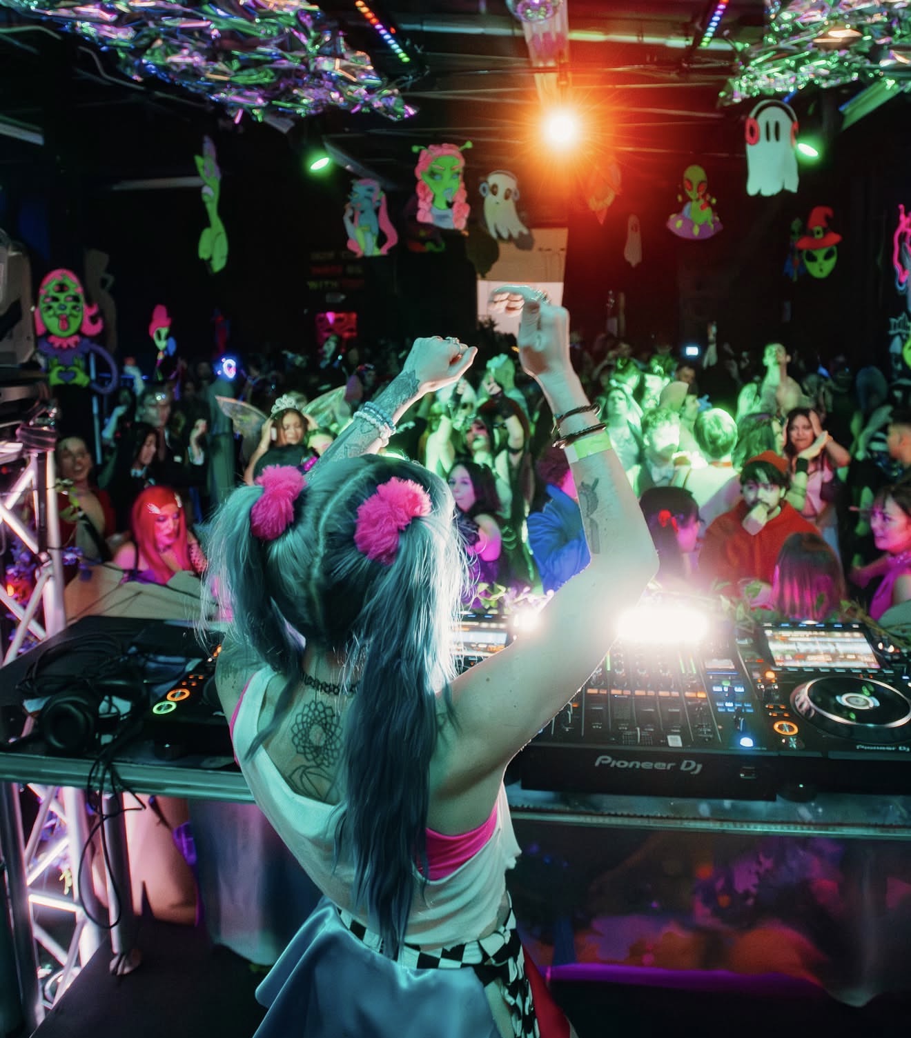 Female DJ with long hair and pink pom-poms, raising her hands at a lively party with colorful decorations and crowd.