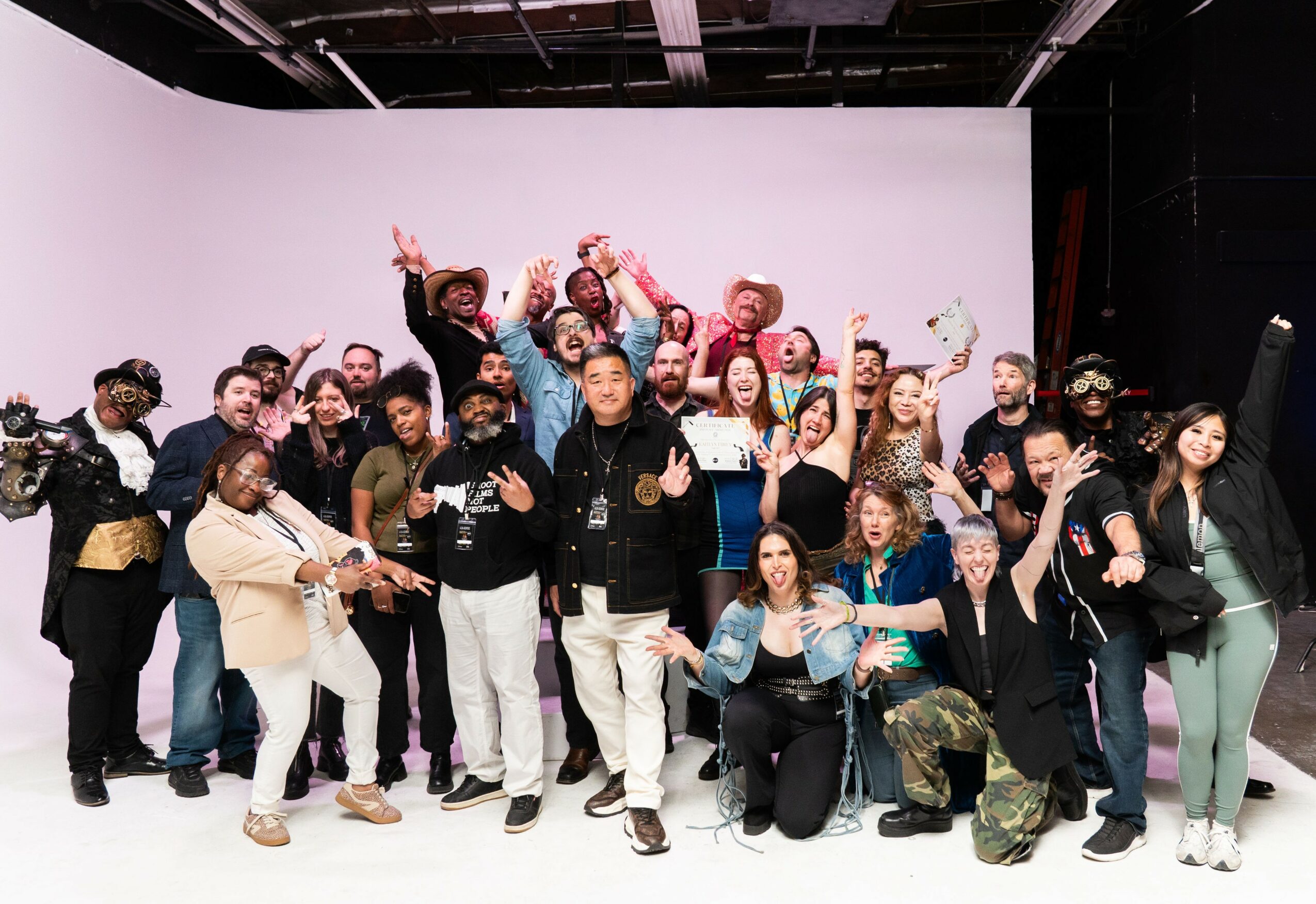 Group of diverse people posing together in a studio with a plain background, some making peace signs and smiling.