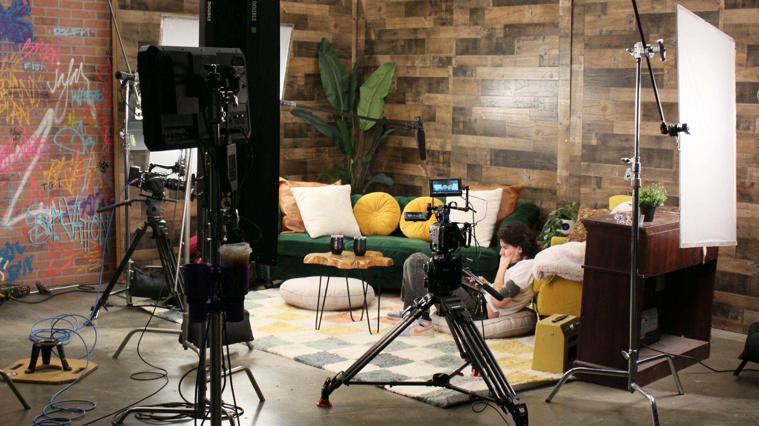 Person lying on a couch with a camera and lighting equipment in a studio setting, wooden wall background, plants, and props.