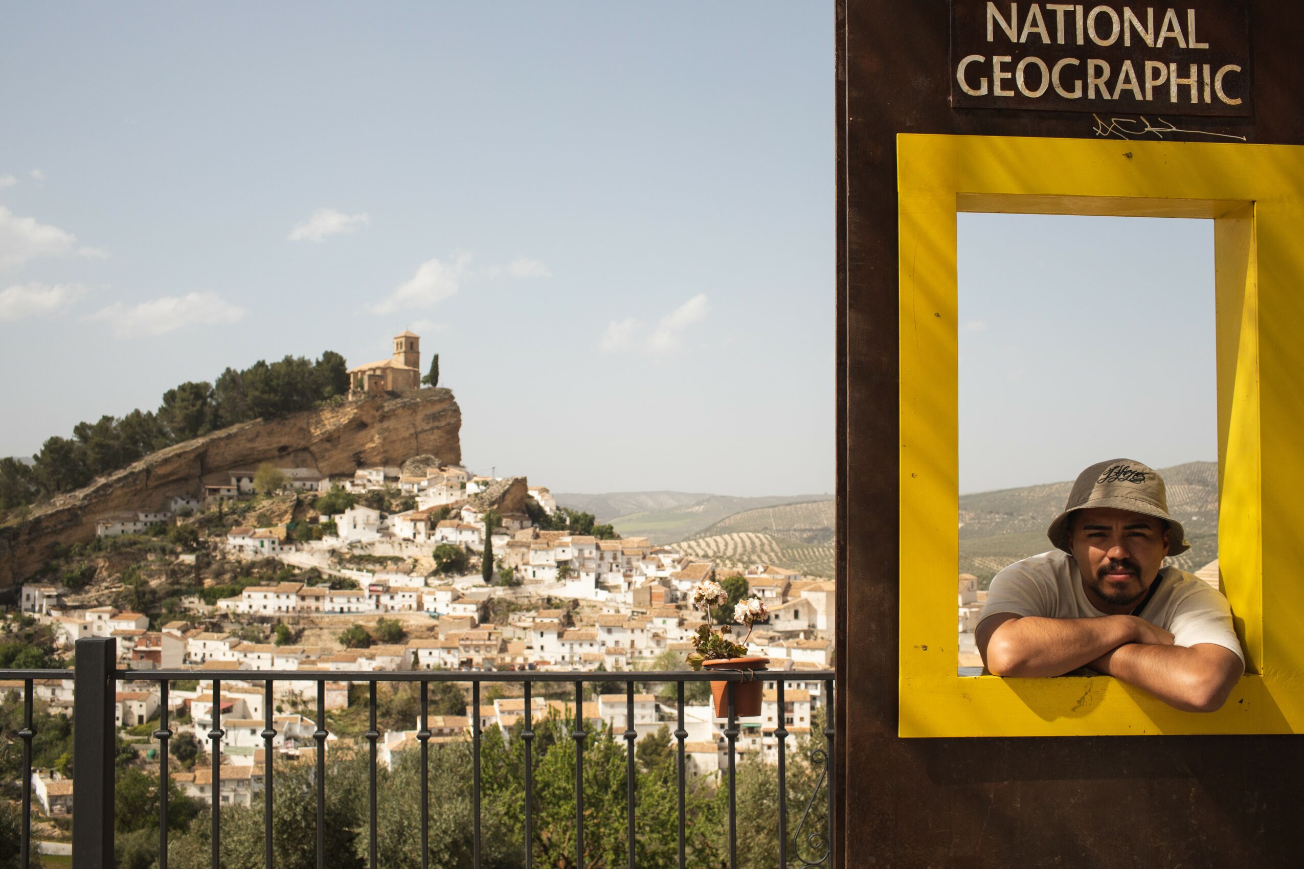 Person with a hat leaning on a yellow frame, overlooking a hillside town with a castle on a hilltop, under a partly cloudy sky.