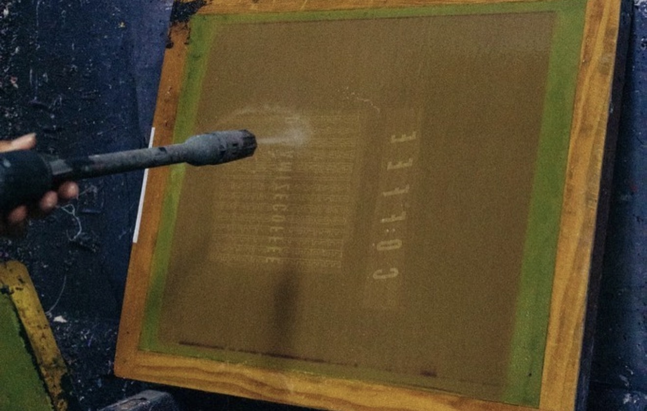 Person cleaning a large, yellow-framed screen printing frame with a spray tool, outdoors in rainy weather.