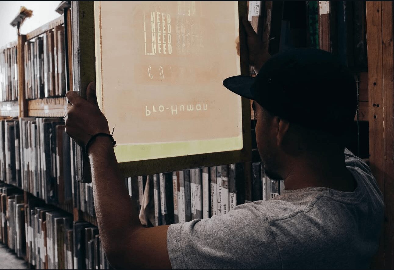 Person adjusting a projector screen in a library with shelves of books, wearing a cap and gray shirt.