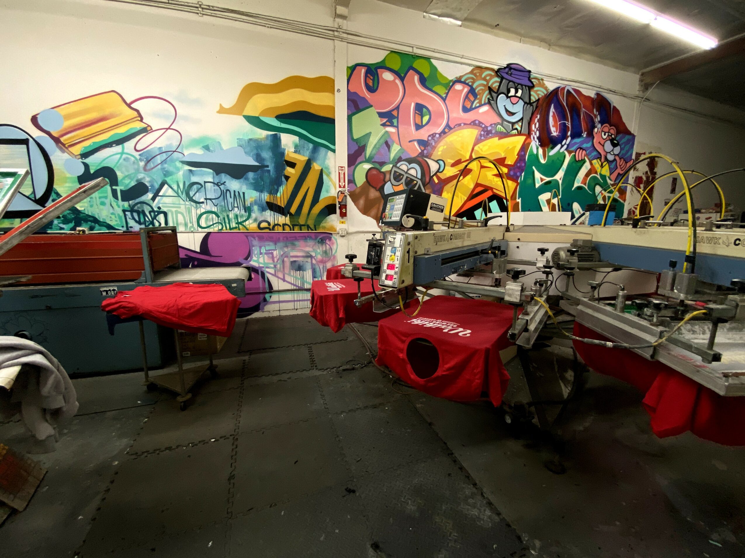 Workshop with graffiti-covered walls, printing machines, and work tables with red cloths, in an industrial setting.