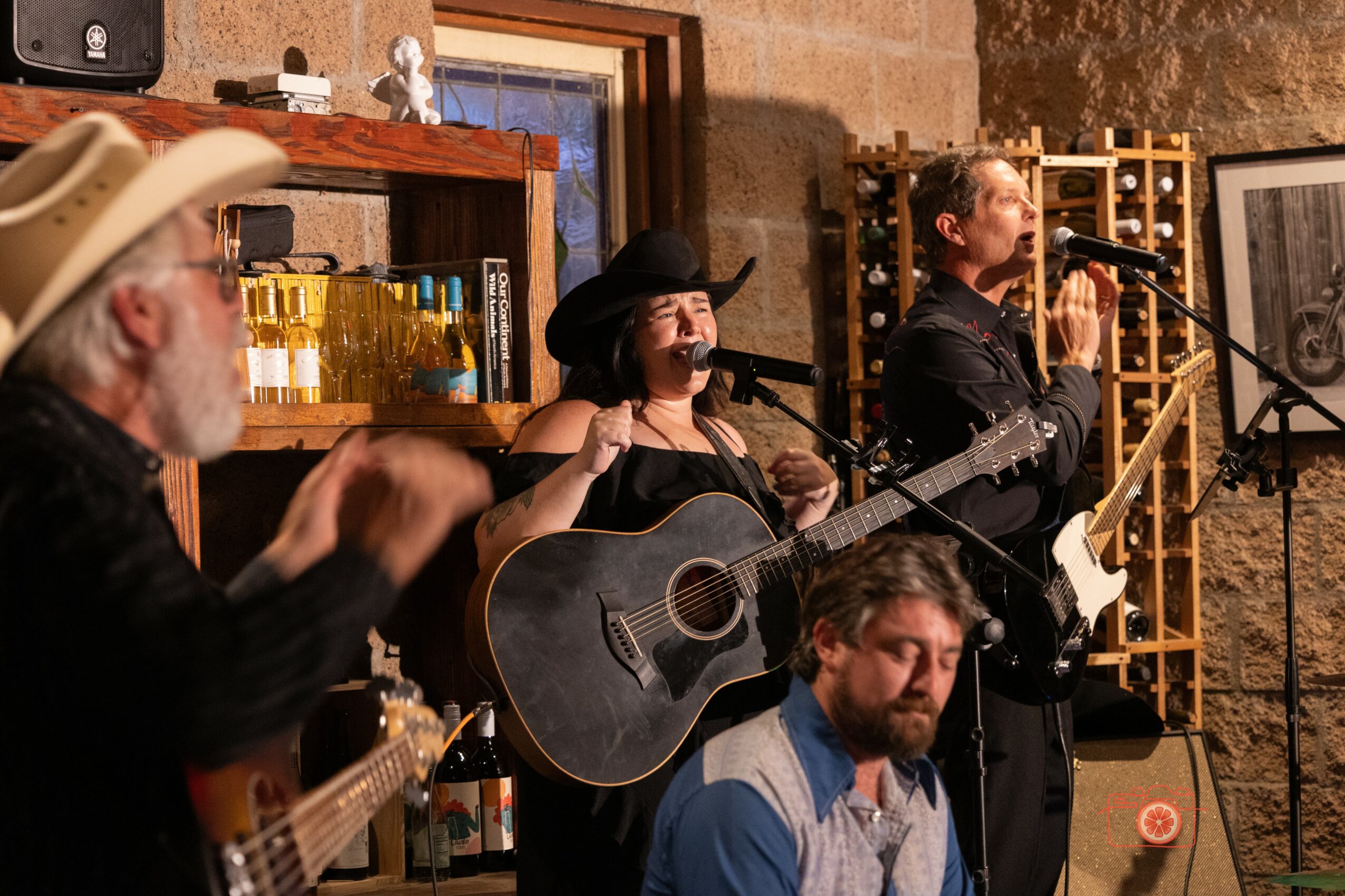 Three musicians perform with guitars and a microphone in a cozy, rustic setting with brick walls and shelves.