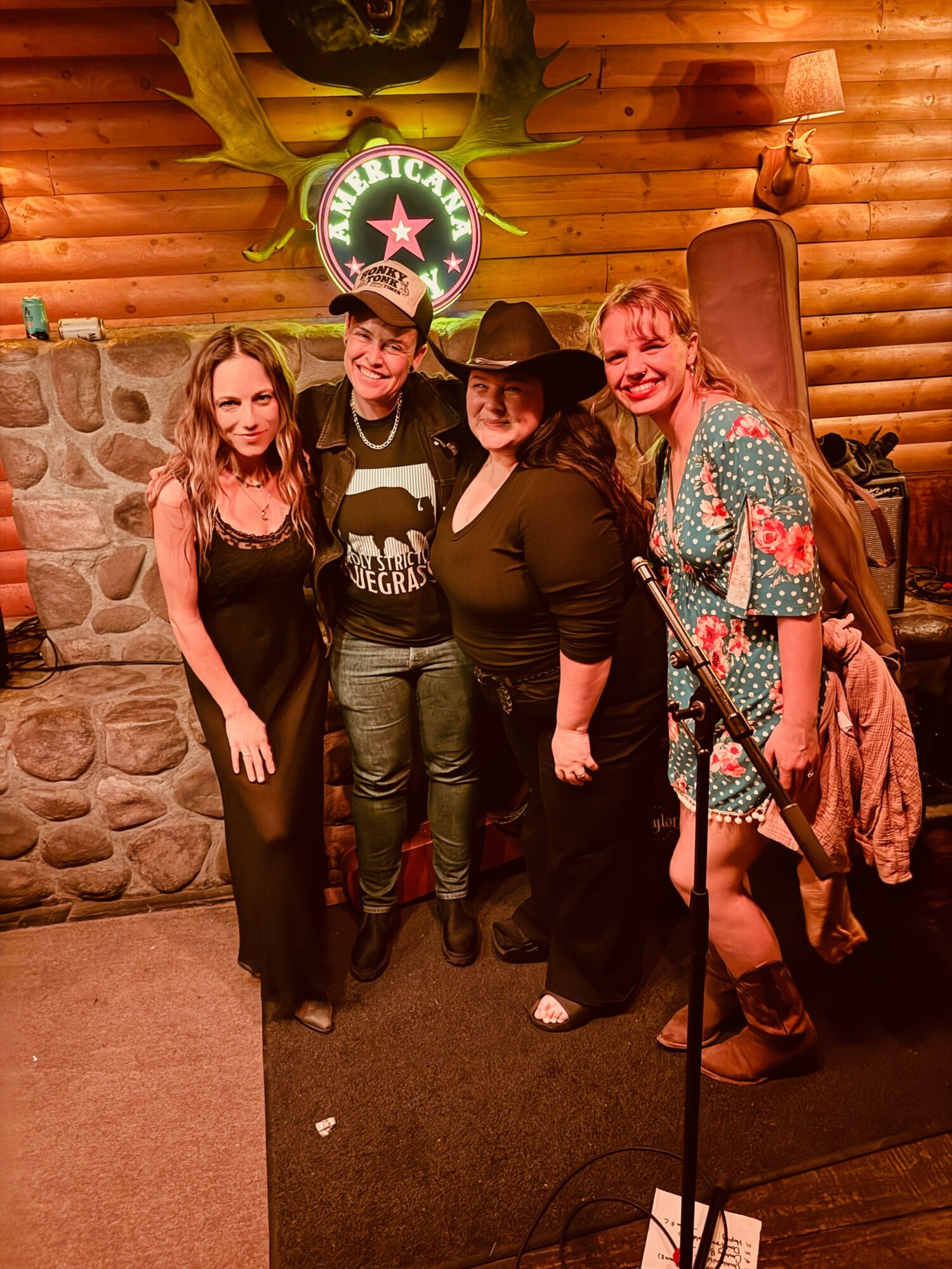 Four women stand together inside a log cabin with a stone fireplace and a neon sign reading 'Welcome.'