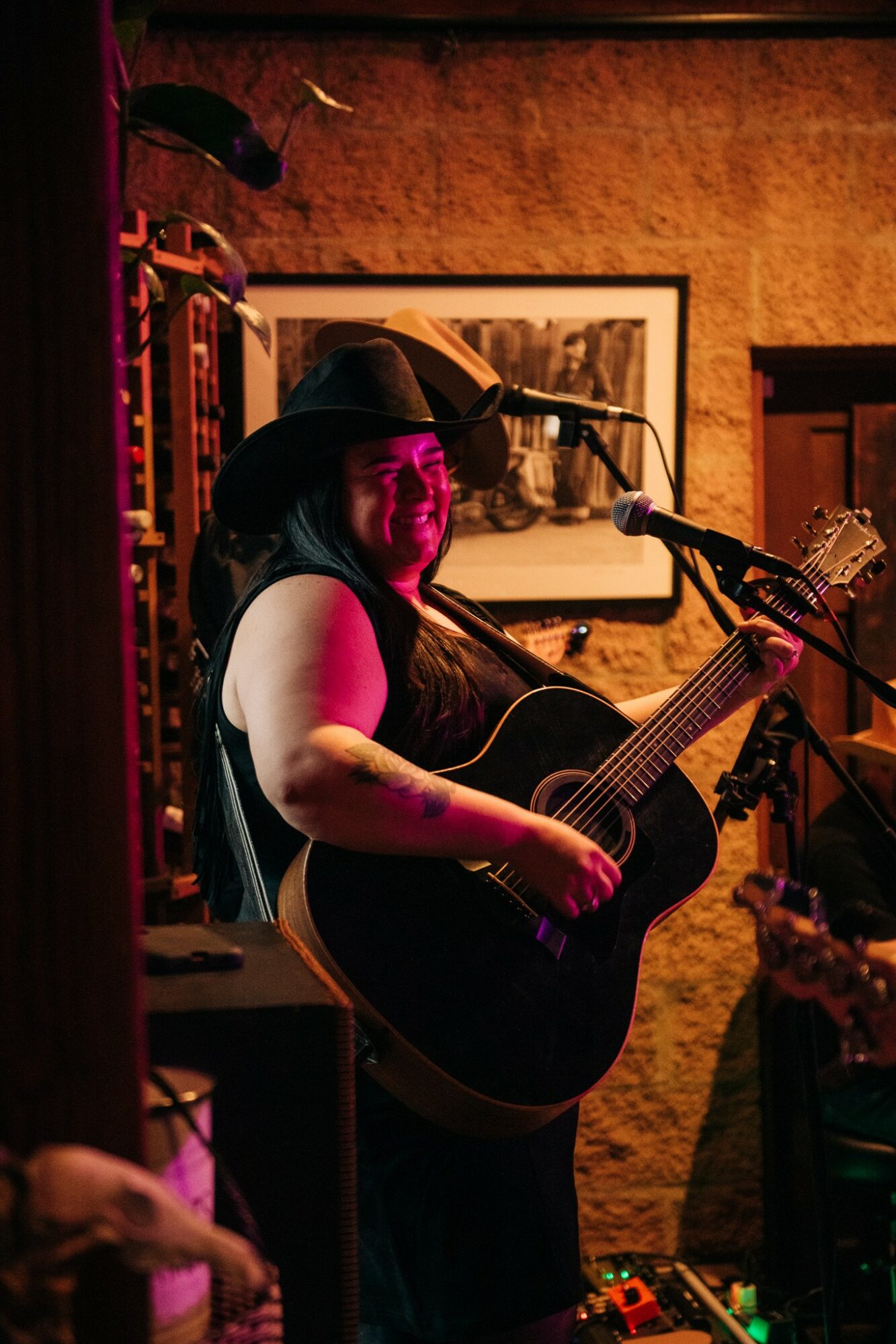 Woman wearing a hat playing an acoustic guitar on stage with warm lighting, framed picture on wall behind her.