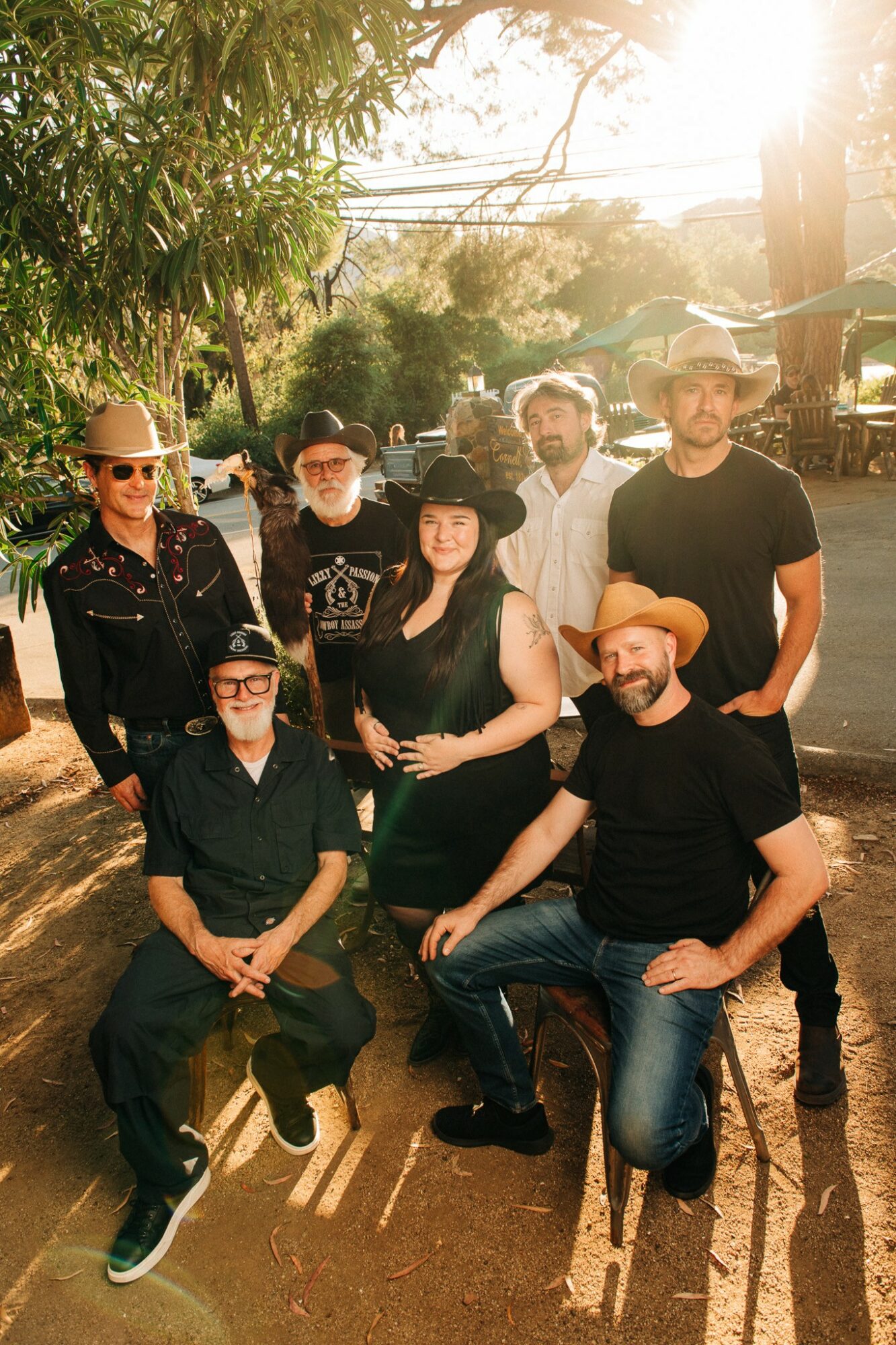 Group of seven people outdoors, some wearing hats and sunglasses, with trees and sunlight in the background.