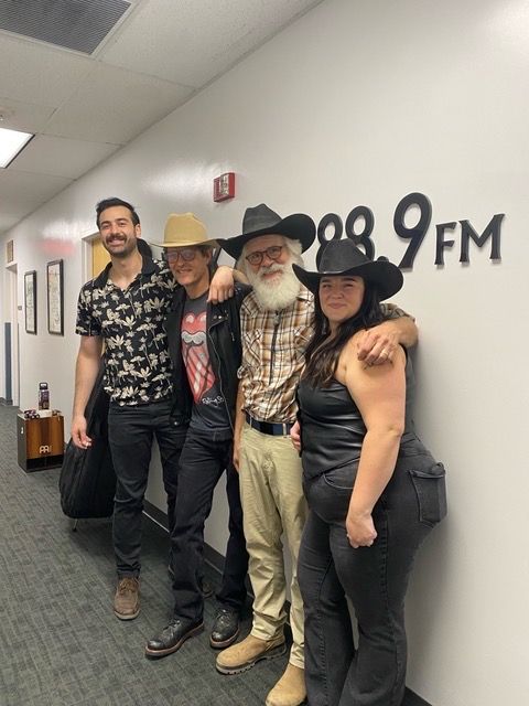 Four people standing in a hallway, one wearing a cowboy hat, with a wall sign reading '90.9 FM'.