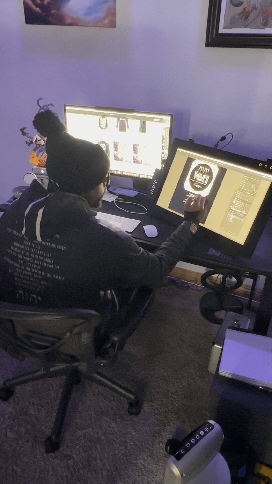 Person working at dual computer monitors in a room with framed pictures on the wall, using a stylus on a graphics tablet.