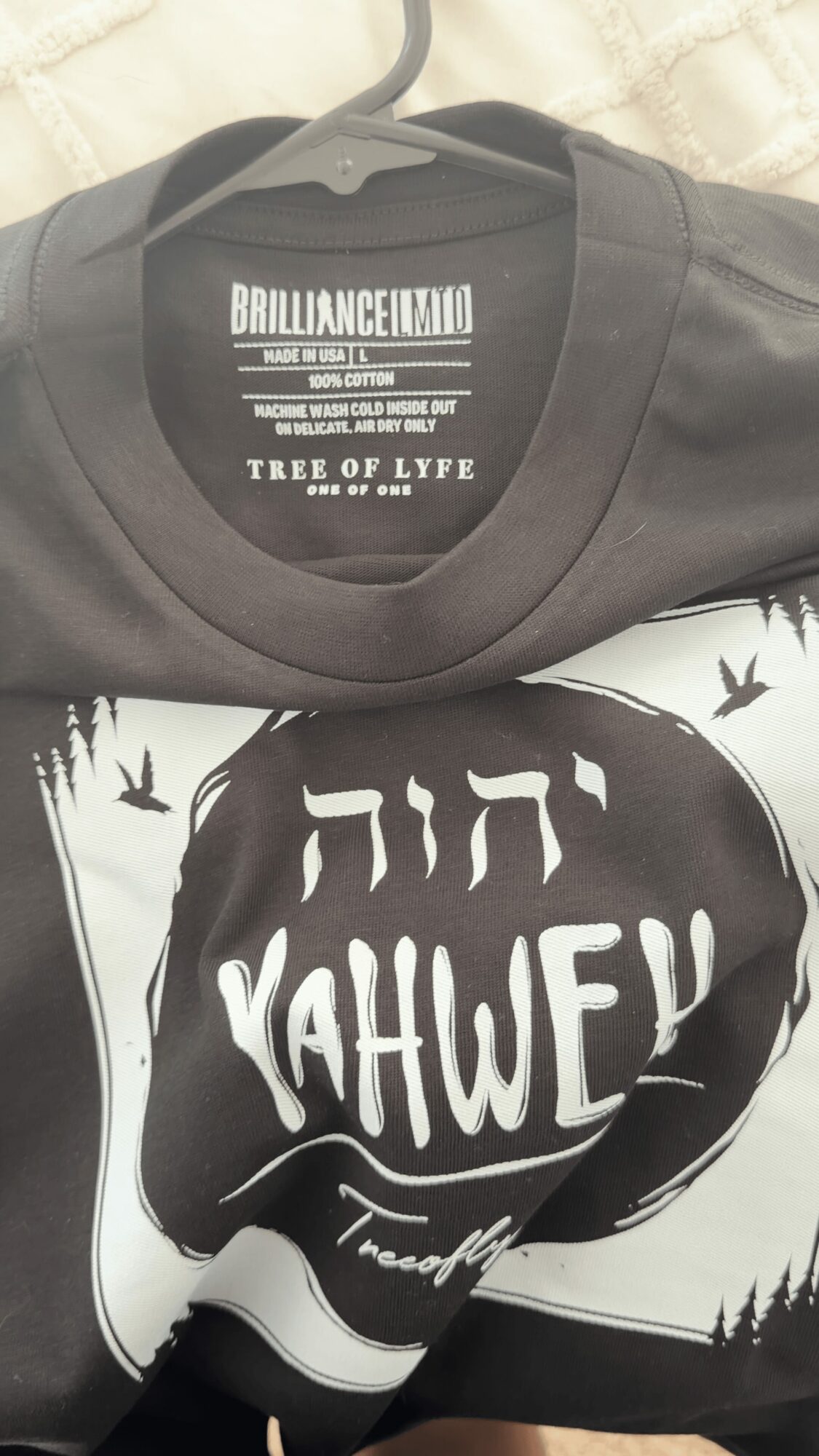Black T-shirt with white text inside a speech bubble, placed on a black and white illustrated background, hanging on a hanger.