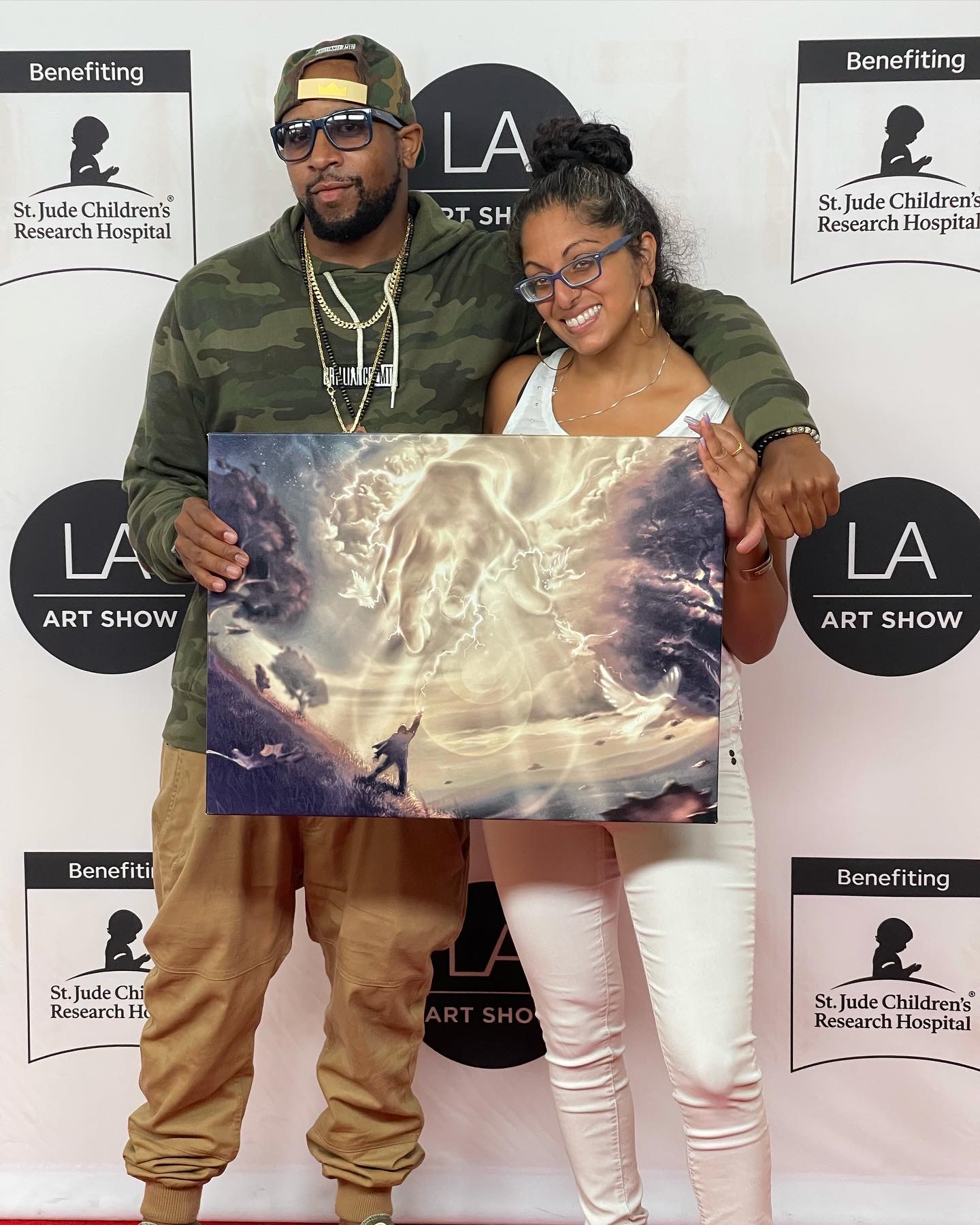 Two people holding a large painting with a cloudy sky and lightning, standing in front of a backdrop with event logos.
