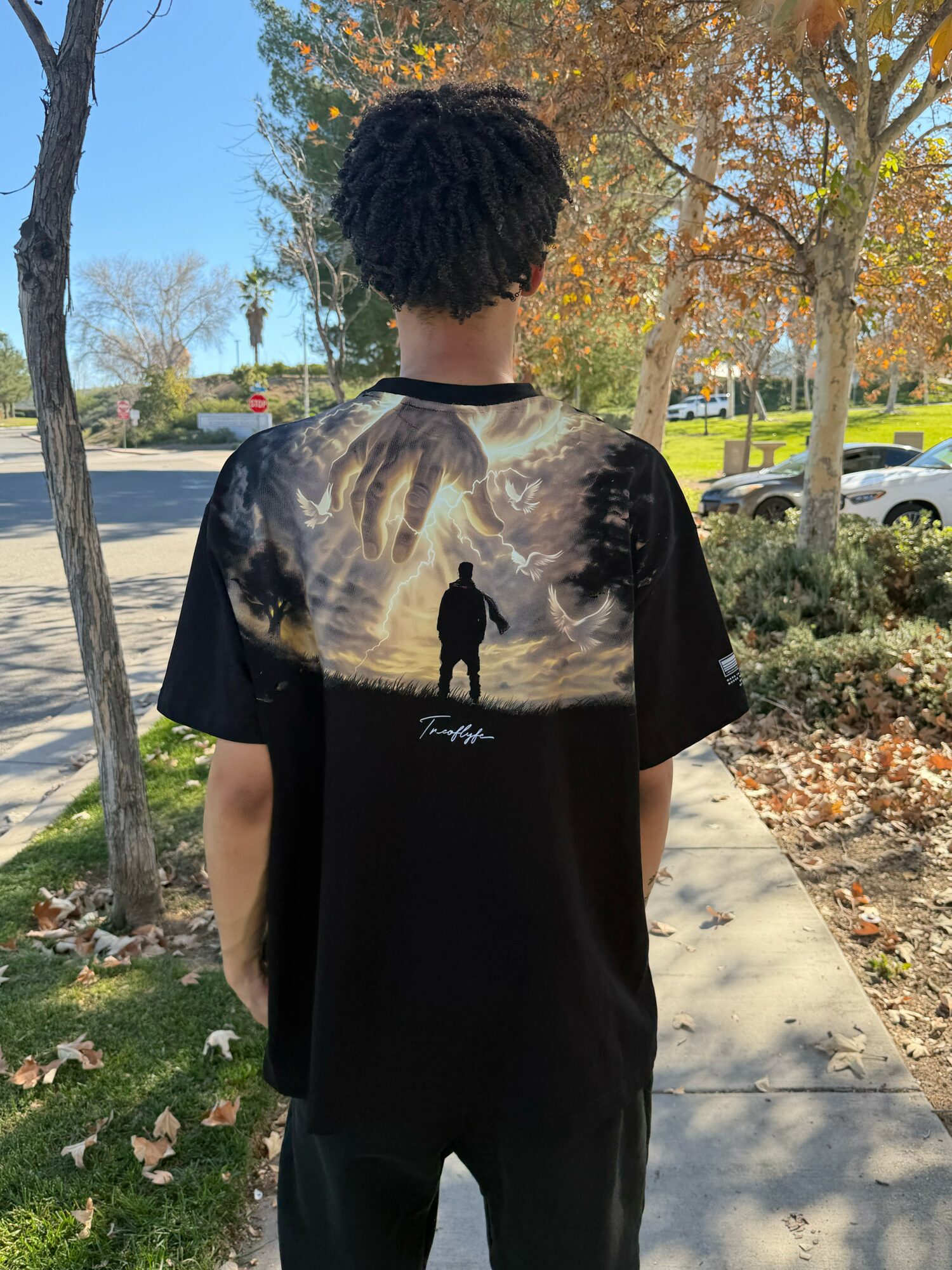 Person with curly hair wearing a black T-shirt with a landscape print on the back, standing outdoors on a sidewalk.