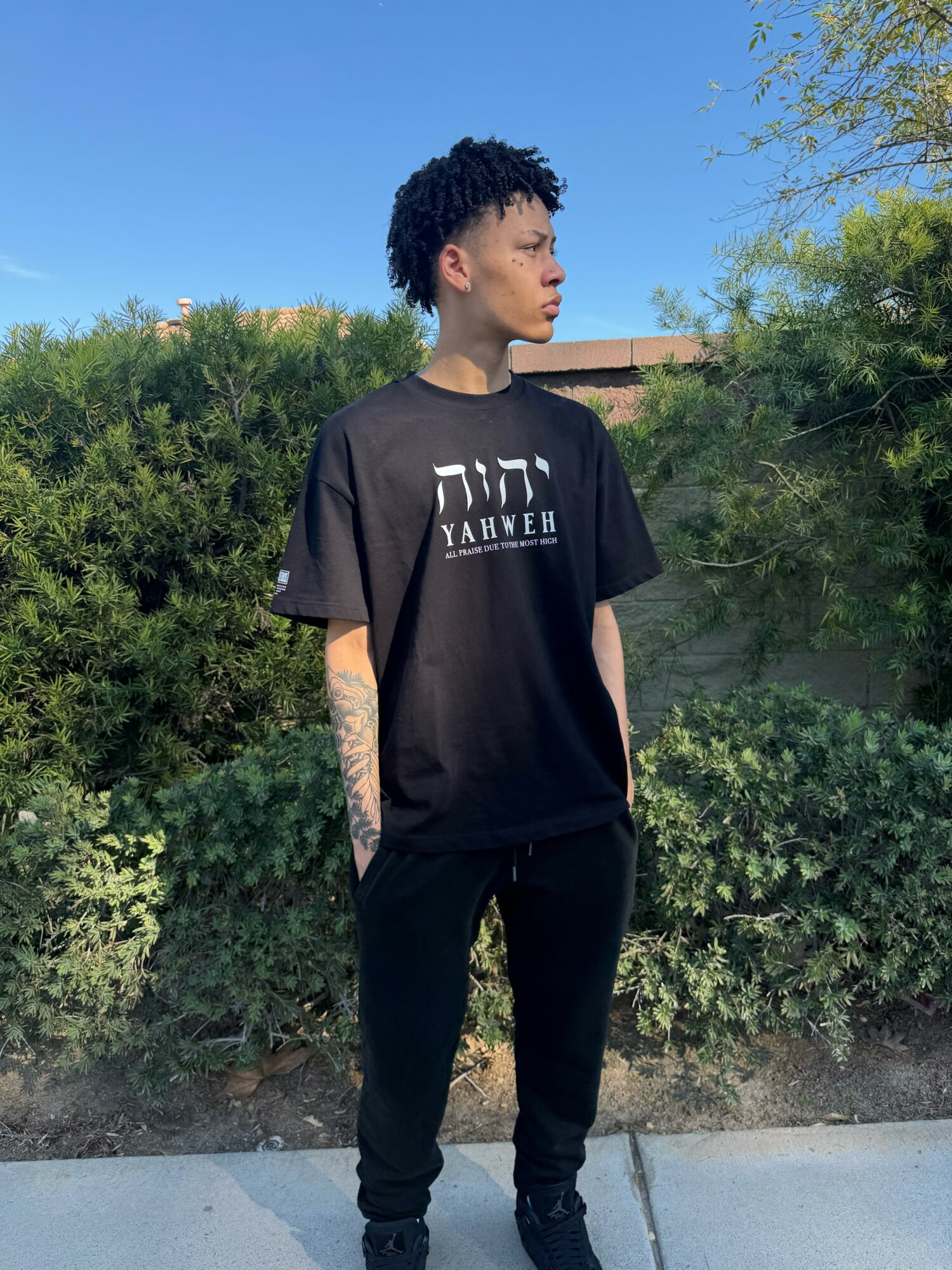 Young person with short curly hair standing outdoors, wearing a black T-shirt and black pants, with greenery and blue sky in background.
