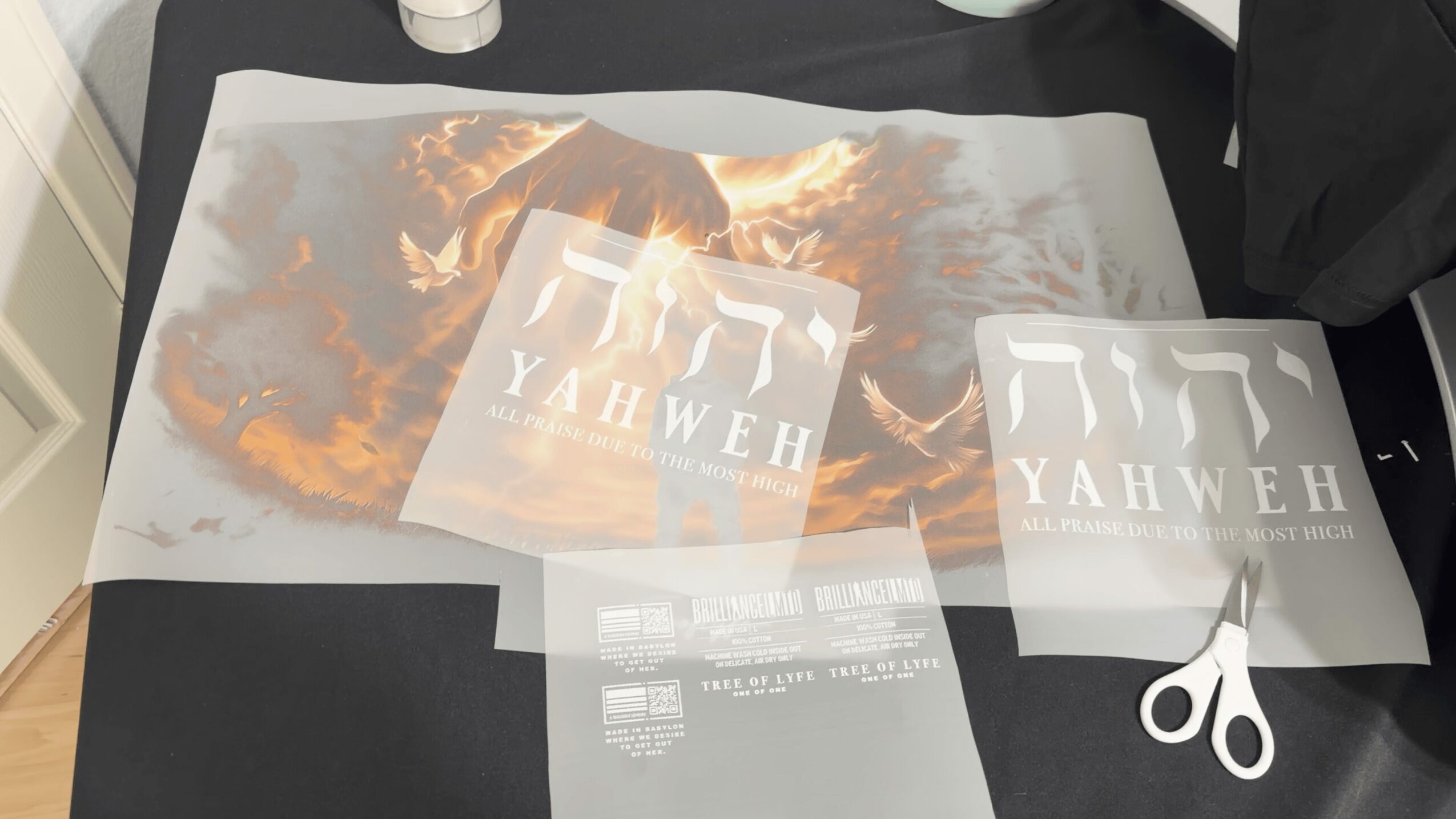 Printed transparent sheet with a fiery background and two logos reading 'YAHWEH' on a black surface, scissors and paper nearby.