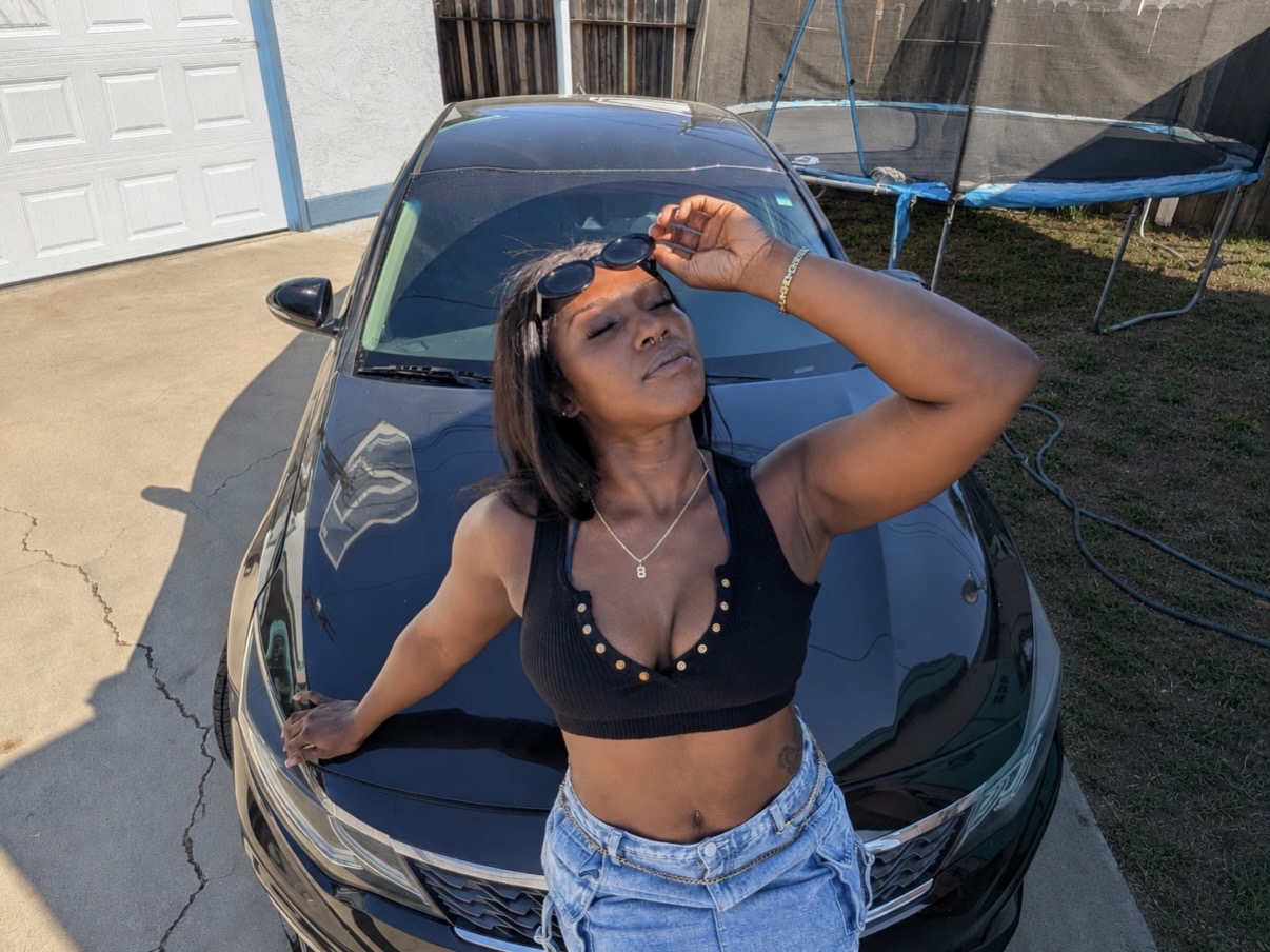 Young woman with short hair, wearing a black crop top and denim shorts, poses outdoors with her hand on her forehead.