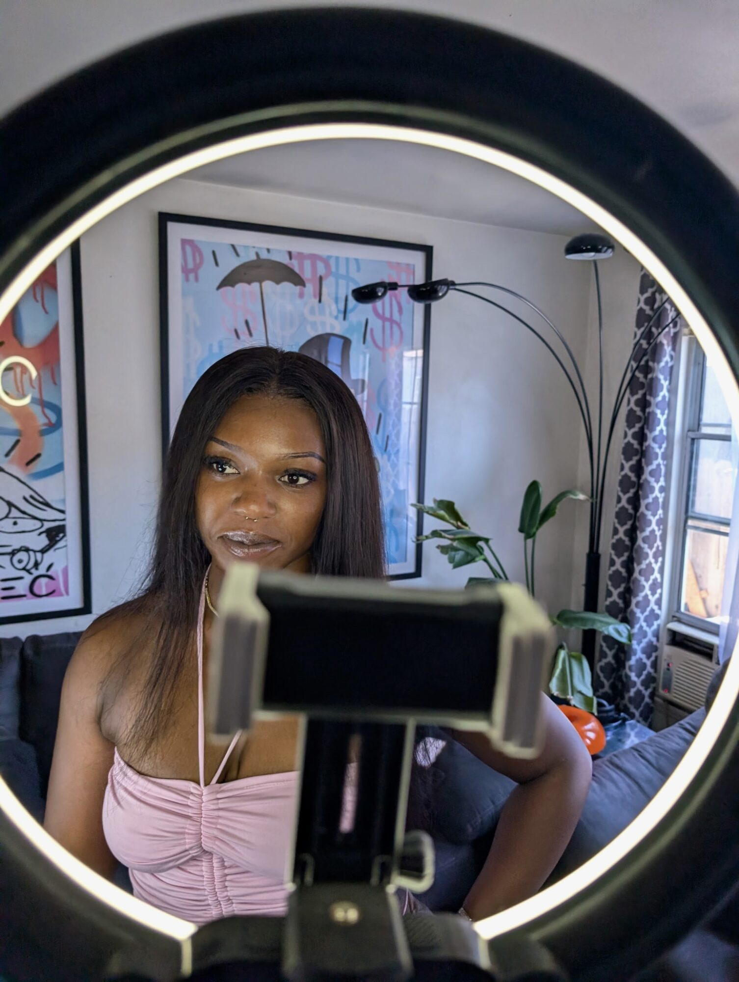 Woman with long dark hair and pink top looking into ring light, mirror reflection, indoor setting with plants and artwork