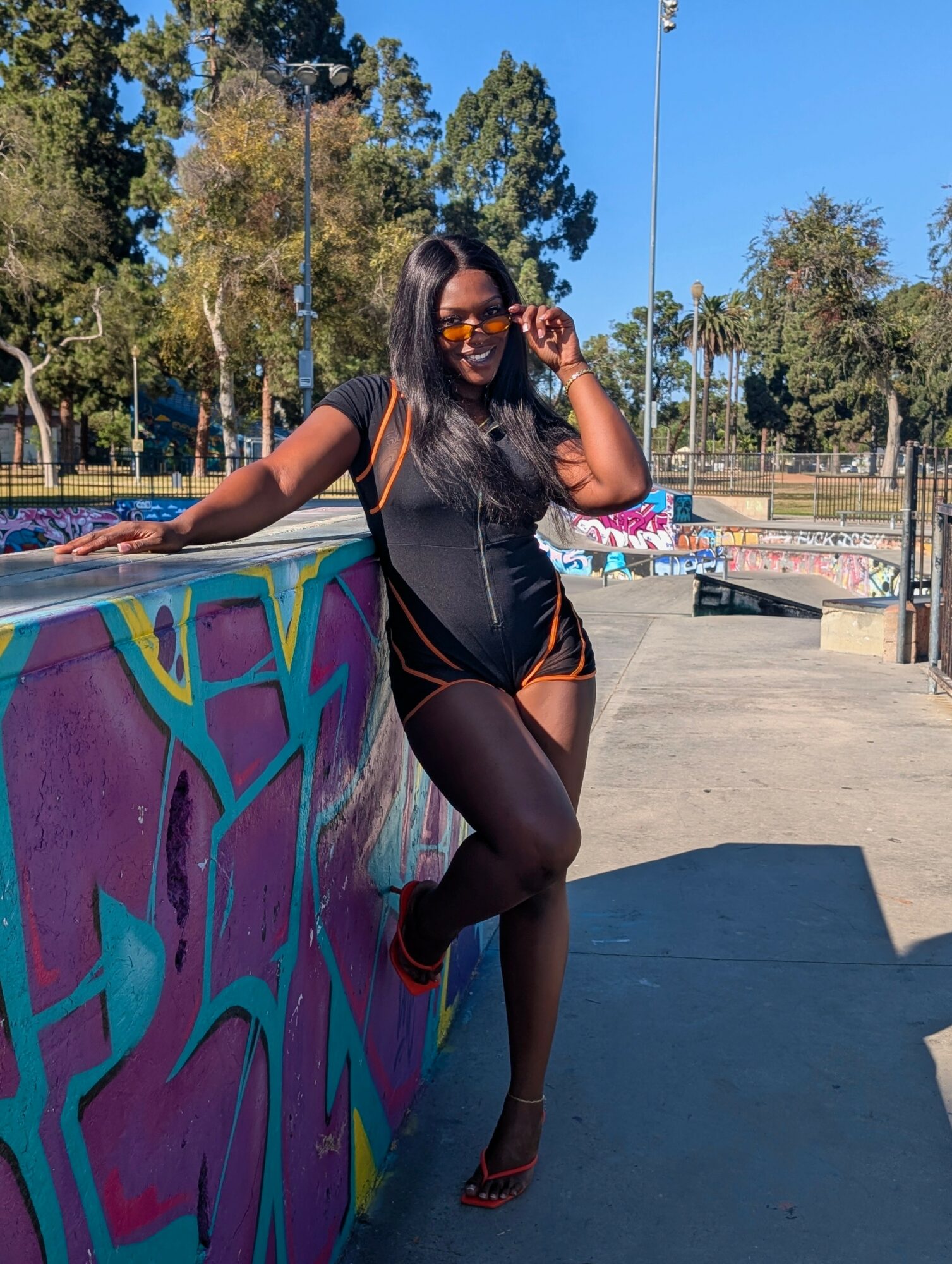Woman with long dark hair wearing sunglasses, black outfit, and heels, leaning on a colorful graffiti wall outdoors.