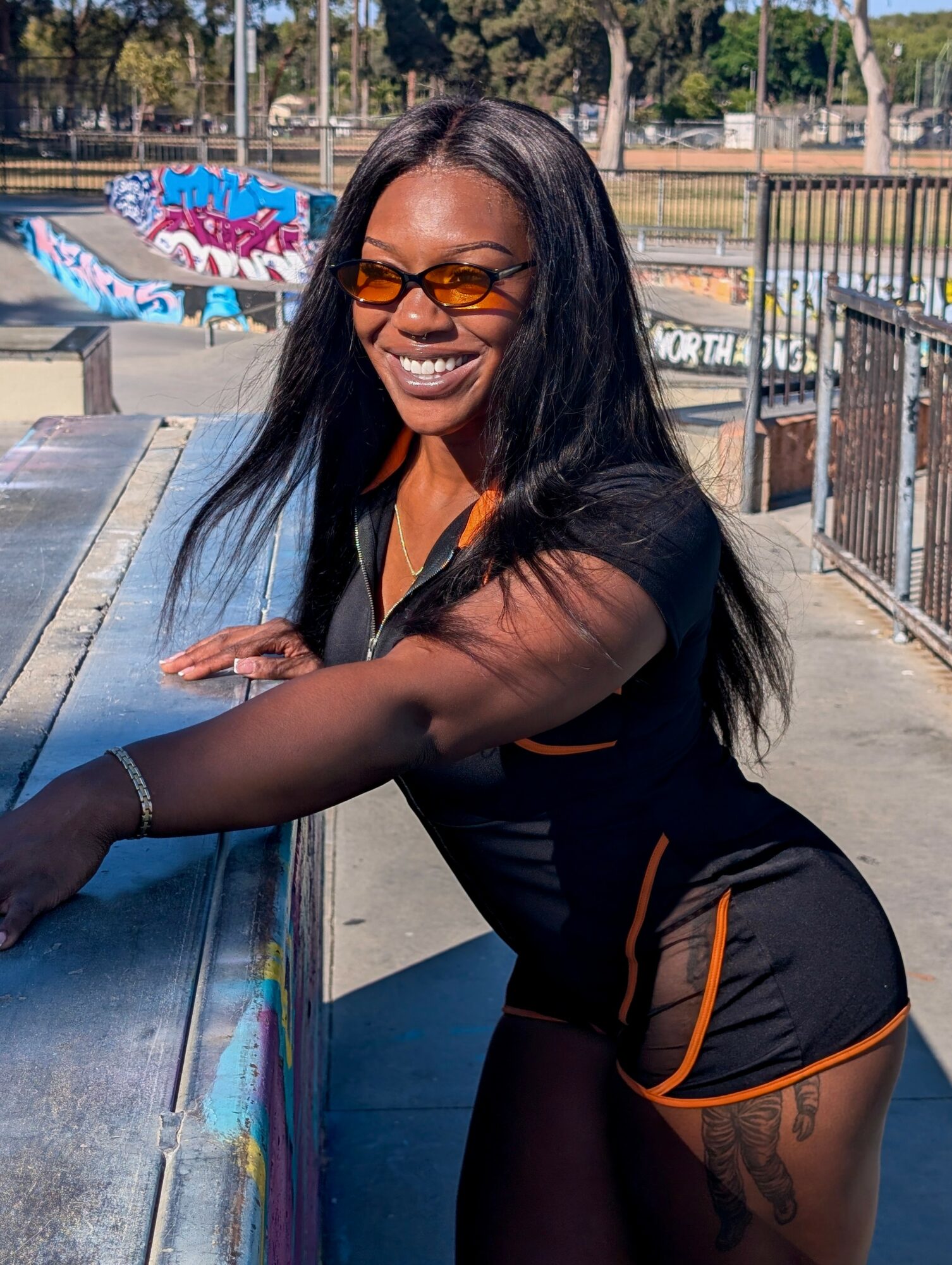 Smiling woman with long dark hair and sunglasses leaning on a railing outdoors, sunny day, colorful graffiti in background.