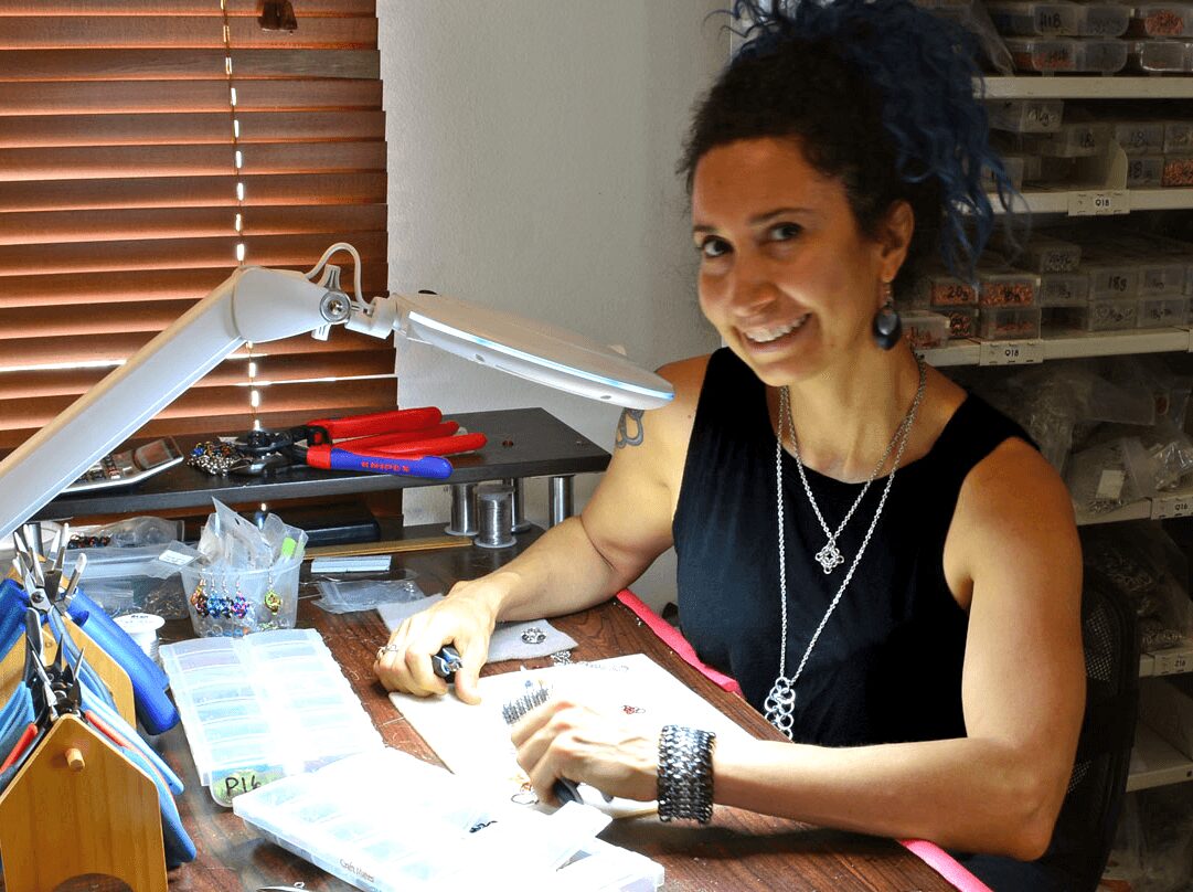 Woman with curly hair smiling at a desk with various supplies, wearing a black sleeveless top and jewelry.