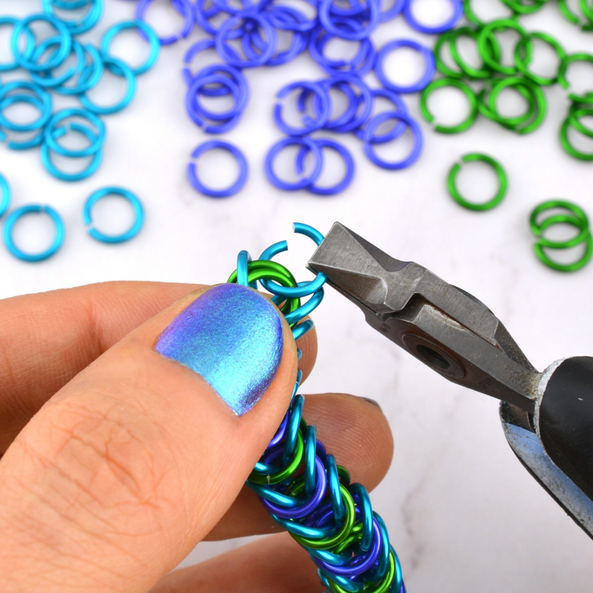 Person's hand with painted fingernail holding colorful metal rings, using pliers to connect rings, with more rings scattered in background.
