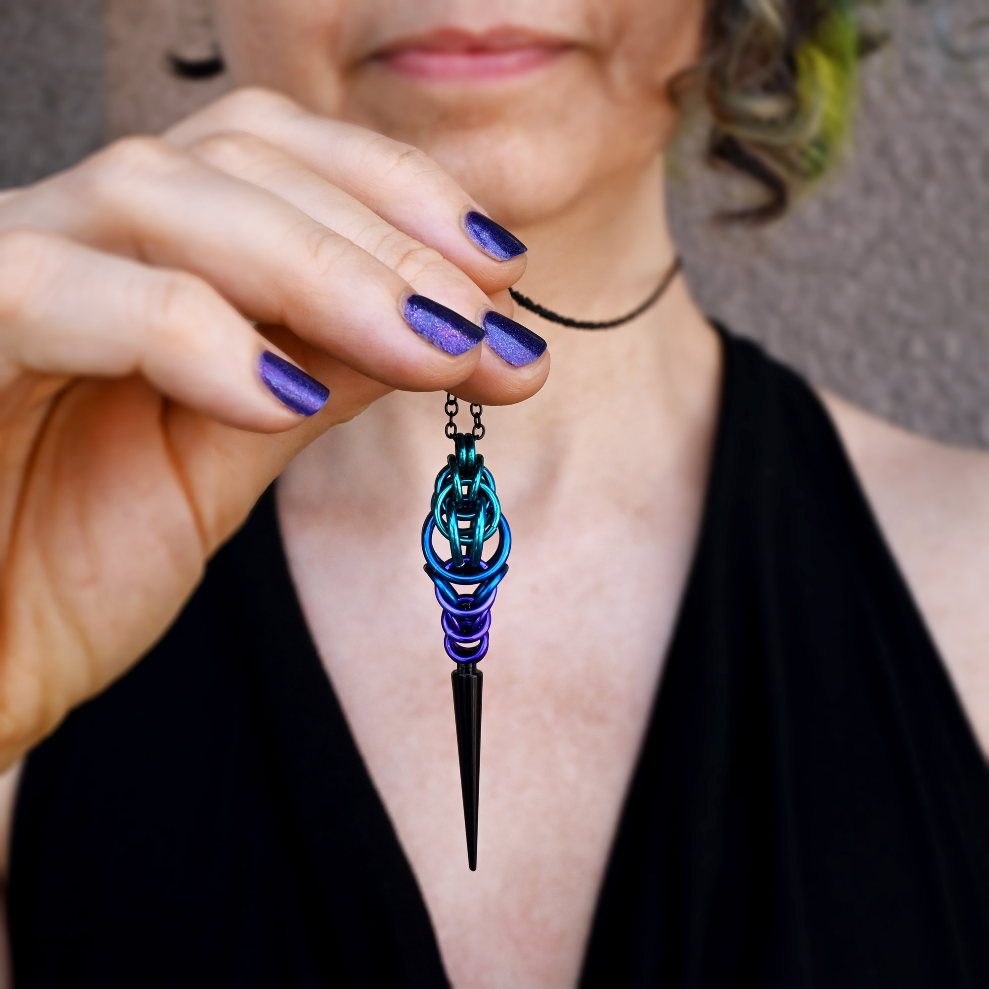 Person holding a blue and black pendant necklace with a pointed tip, wearing a black top and choker, against a gray background.