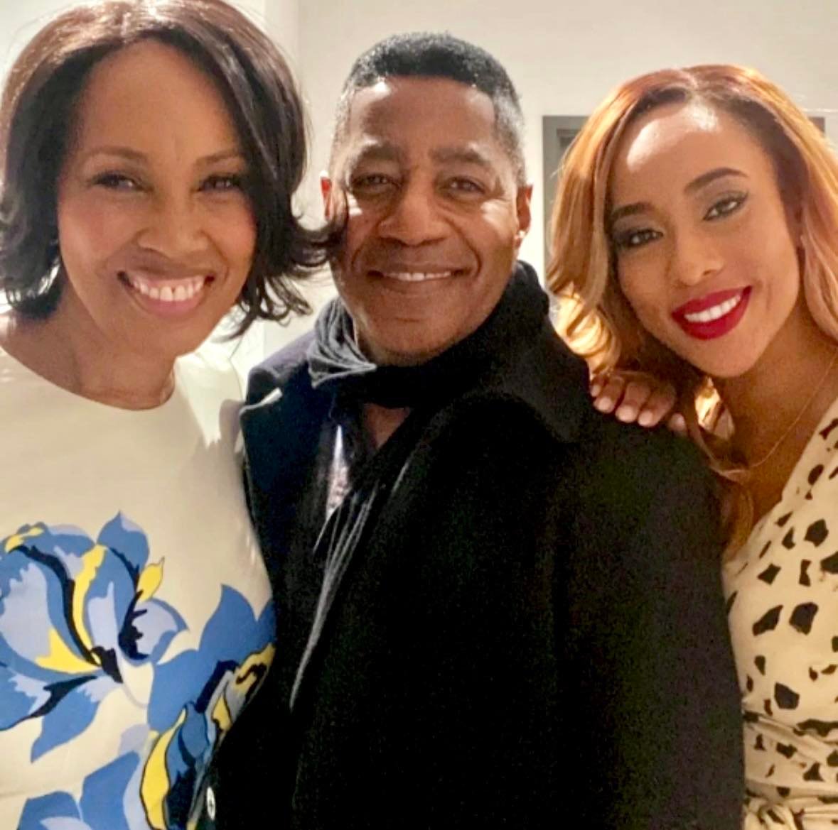 Three people smiling closely, two women and one man, indoors with neutral background.