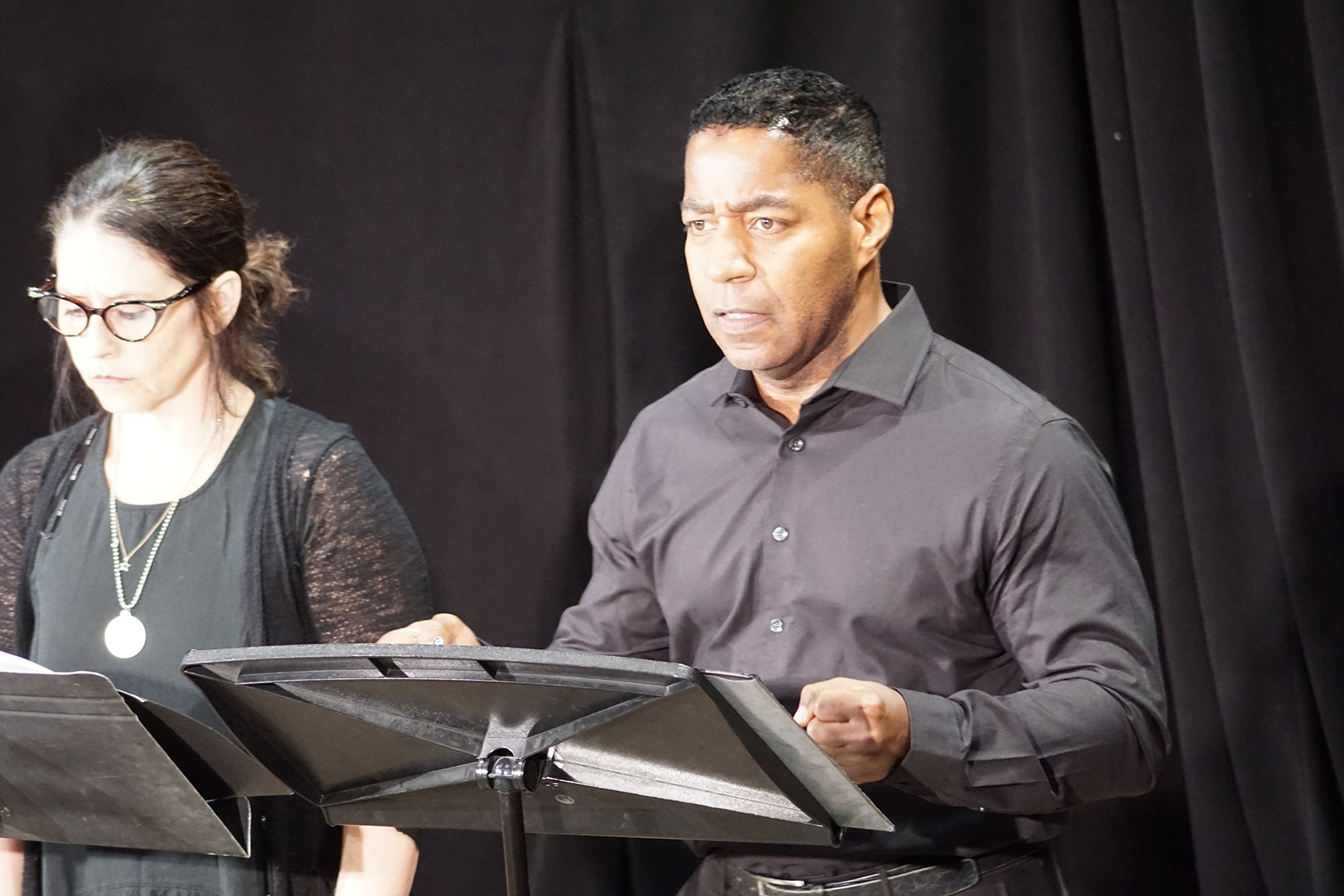 Two people stand behind music stands, one man and one woman, in front of a black curtain, focused on their music.