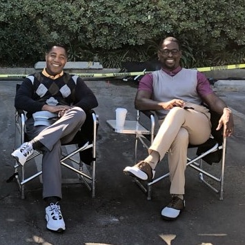 Two men sitting on chairs outdoors, smiling, with trees in the background and a yellow caution tape behind them.