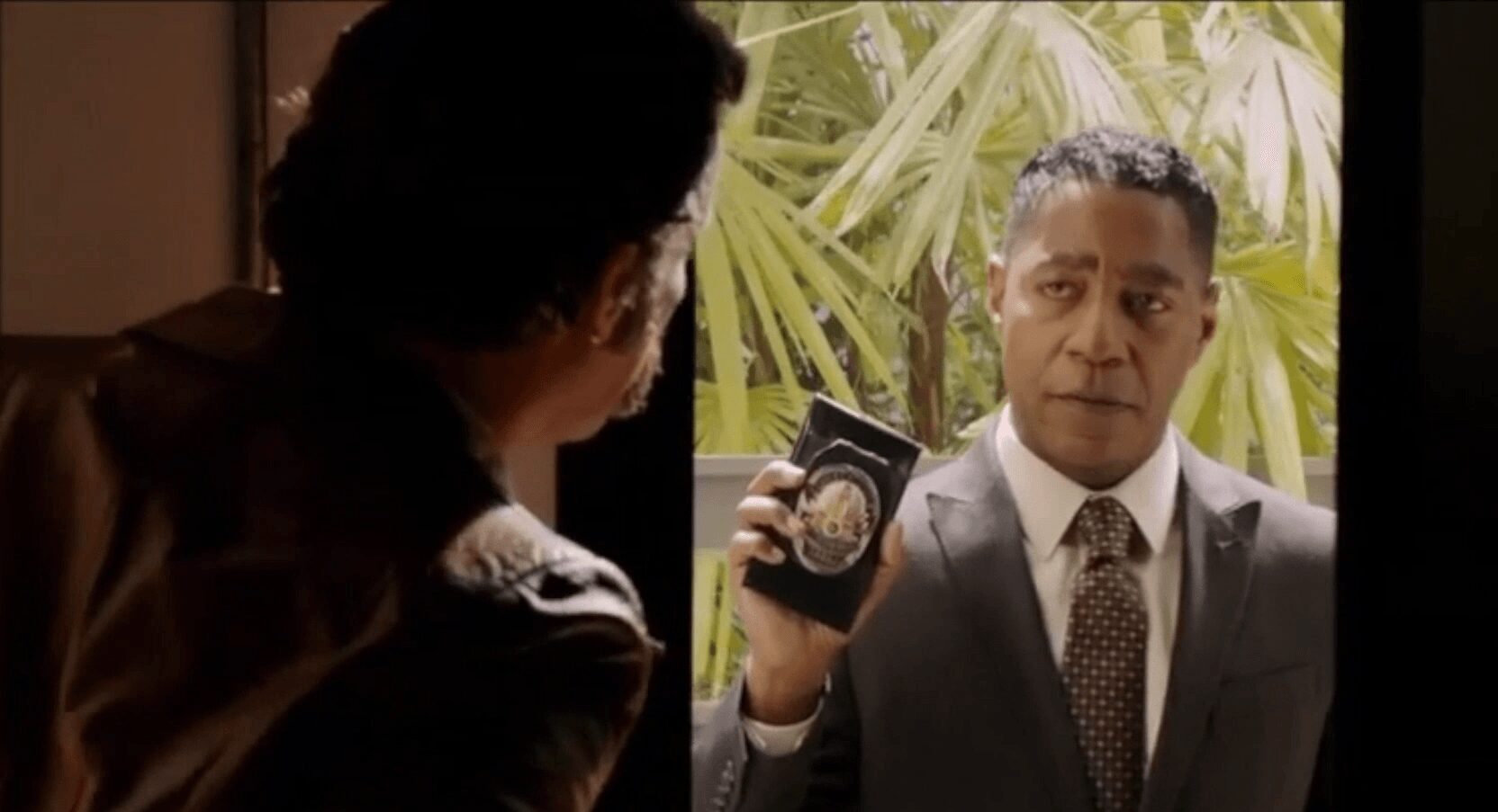 Two men face each other, one holding a badge, outside with large green plants in the background.