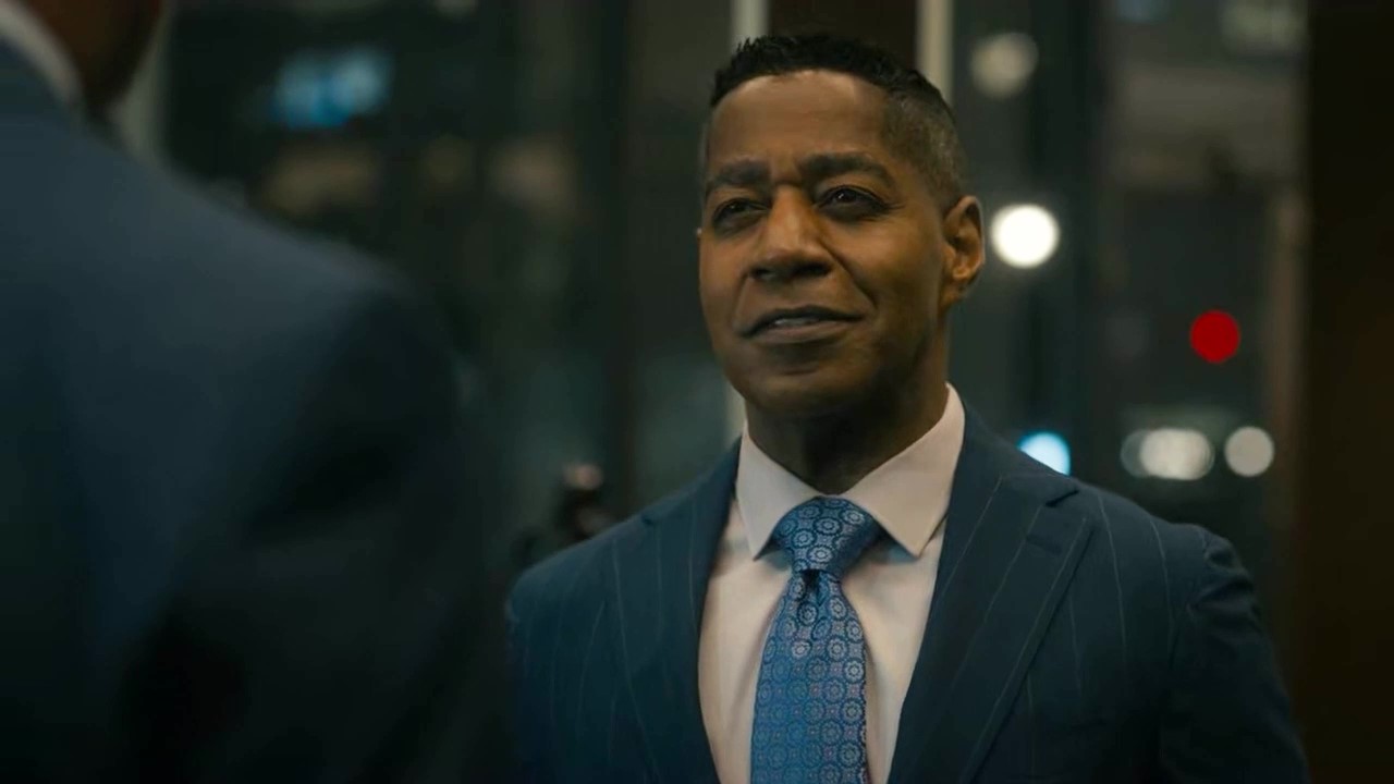 Man in a suit and tie standing indoors, facing another person, with city lights in the background.