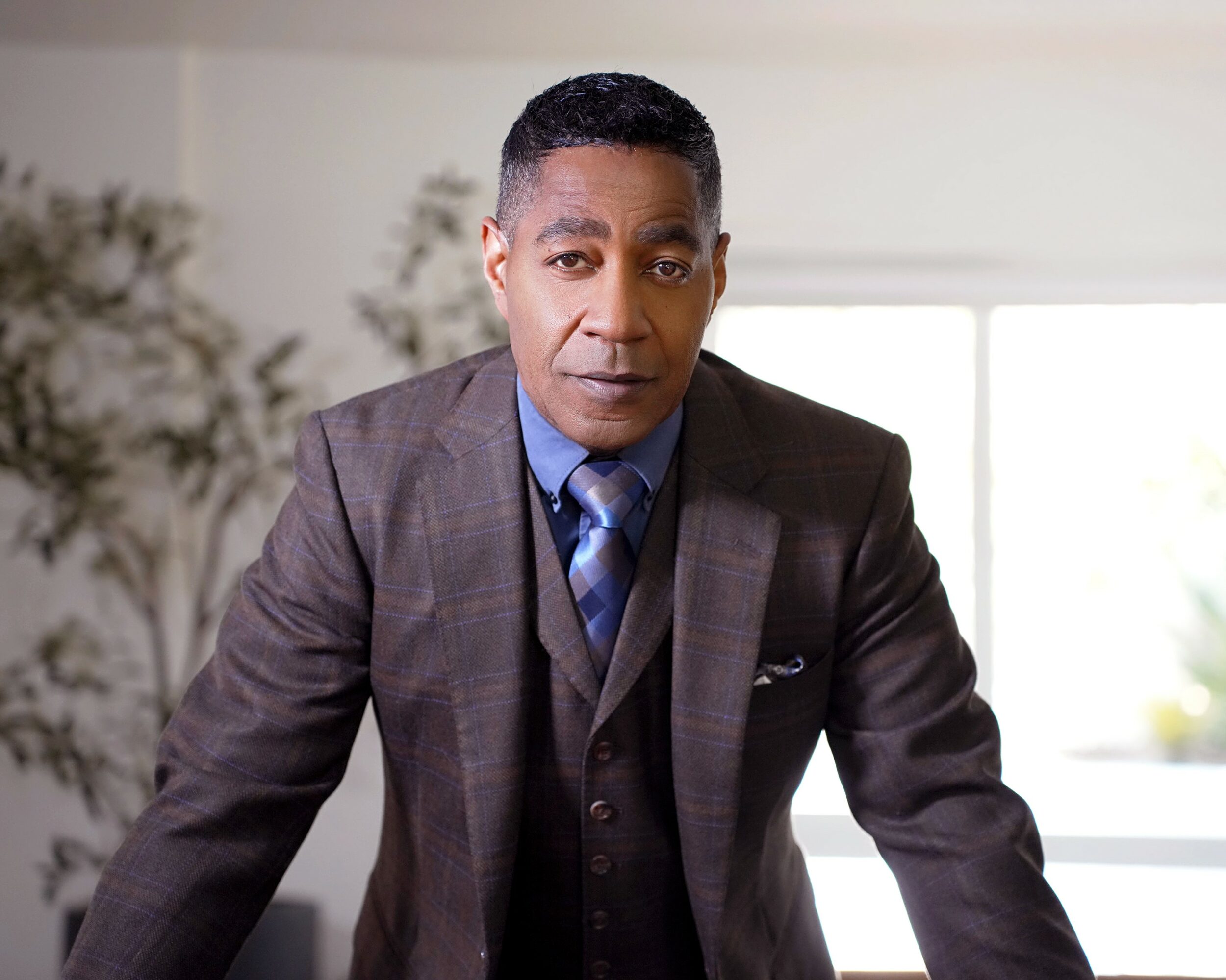 Man in a suit leaning forward indoors with plants and a window in the background.