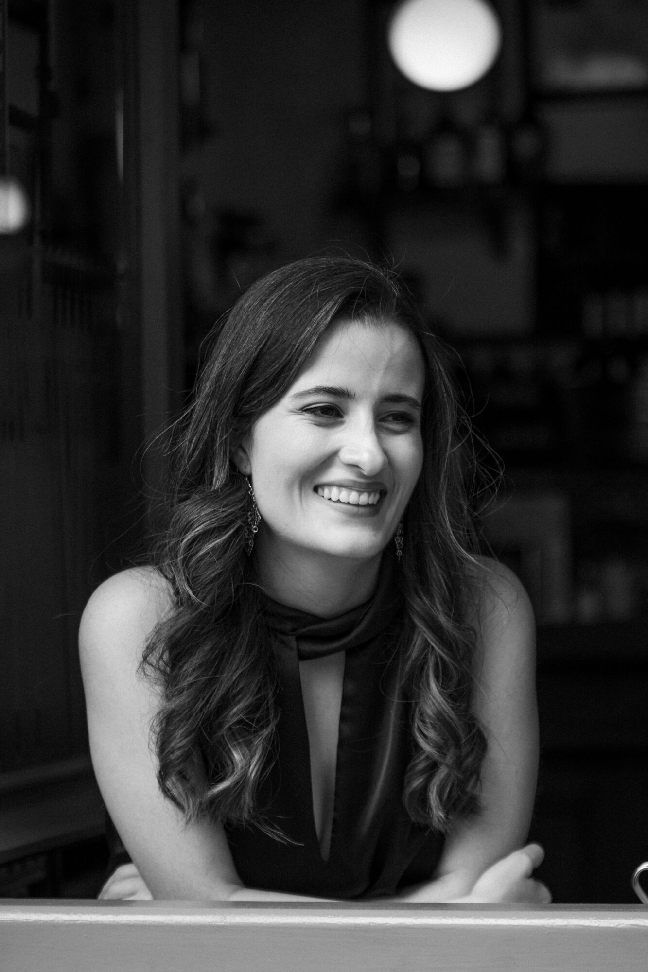 Smiling woman with long wavy hair sitting indoors, background blurred, black and white photo.