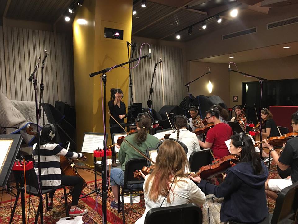Orchestra rehearsal with musicians playing string instruments in a studio with microphones and music stands.