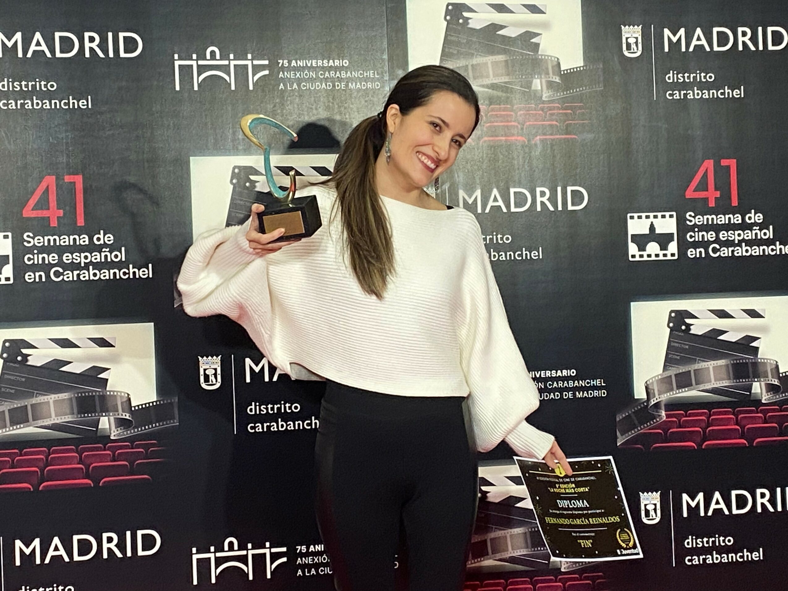 Woman smiling holding an award and a certificate in front of a Madrid cinema backdrop.