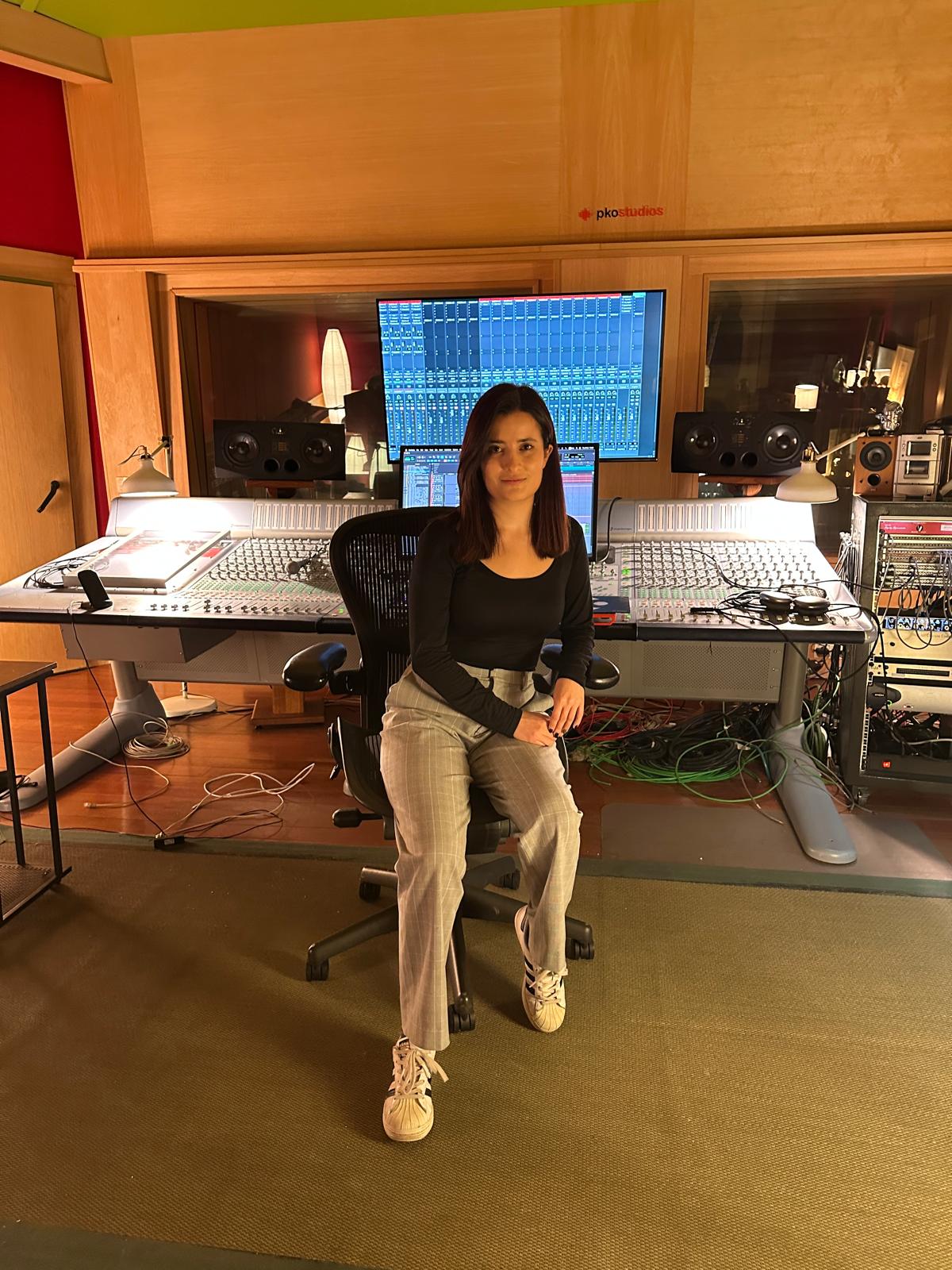 Woman sitting in a recording studio with audio equipment and a large monitor behind her.