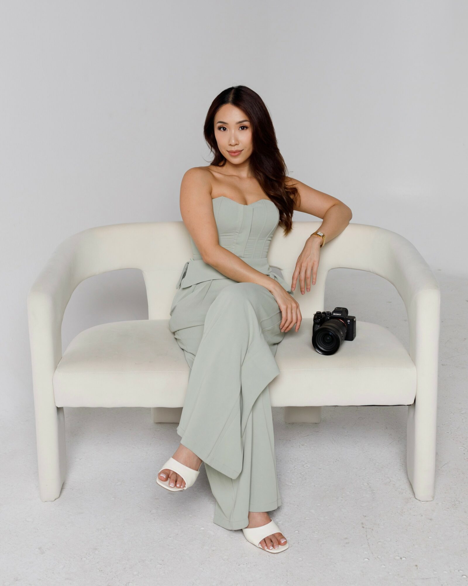 Woman sitting on a white sofa with a camera beside her, wearing a light-colored strapless outfit and white sandals.