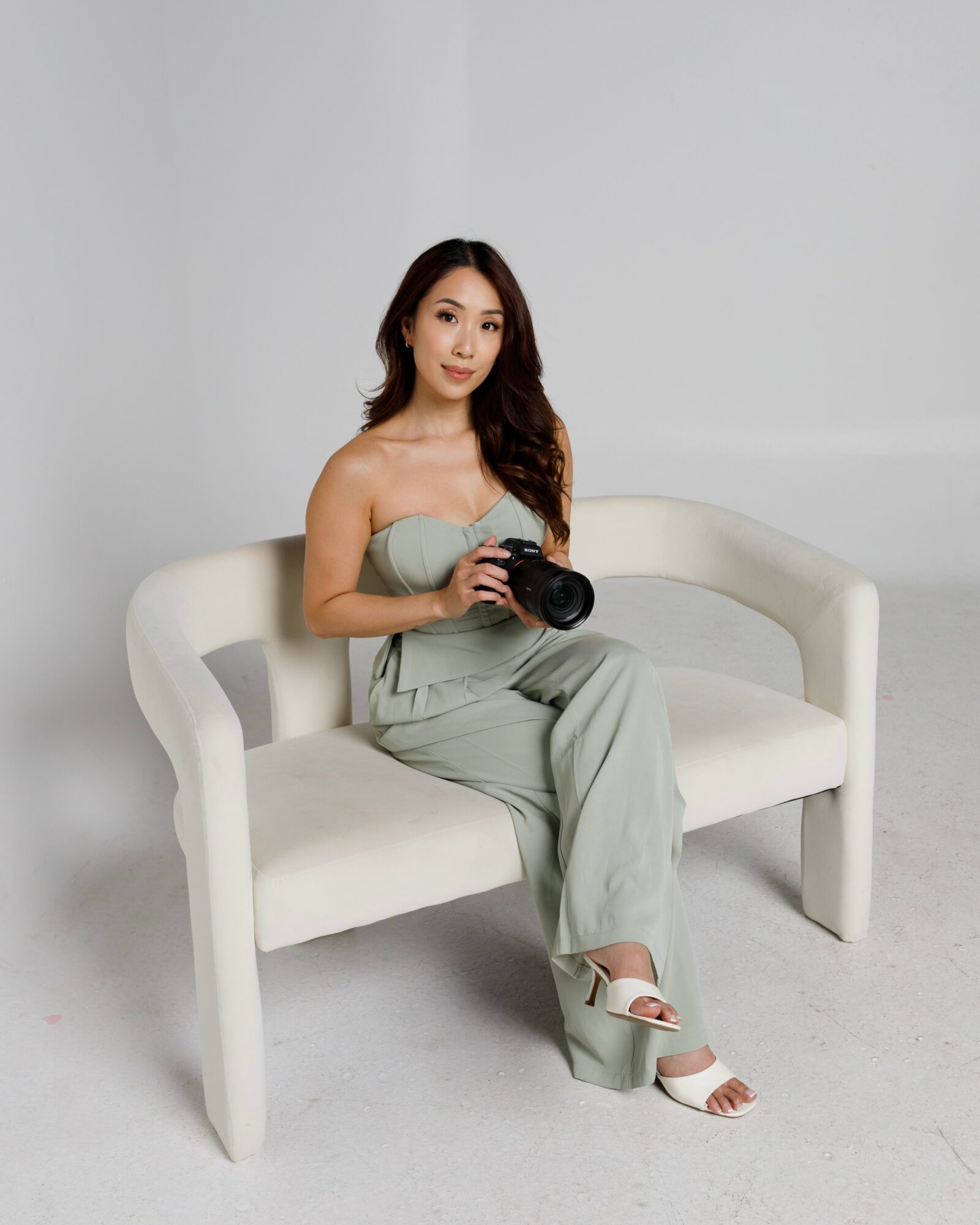Woman sitting on a white bench holding a camera, wearing a light-colored strapless jumpsuit and white sandals.