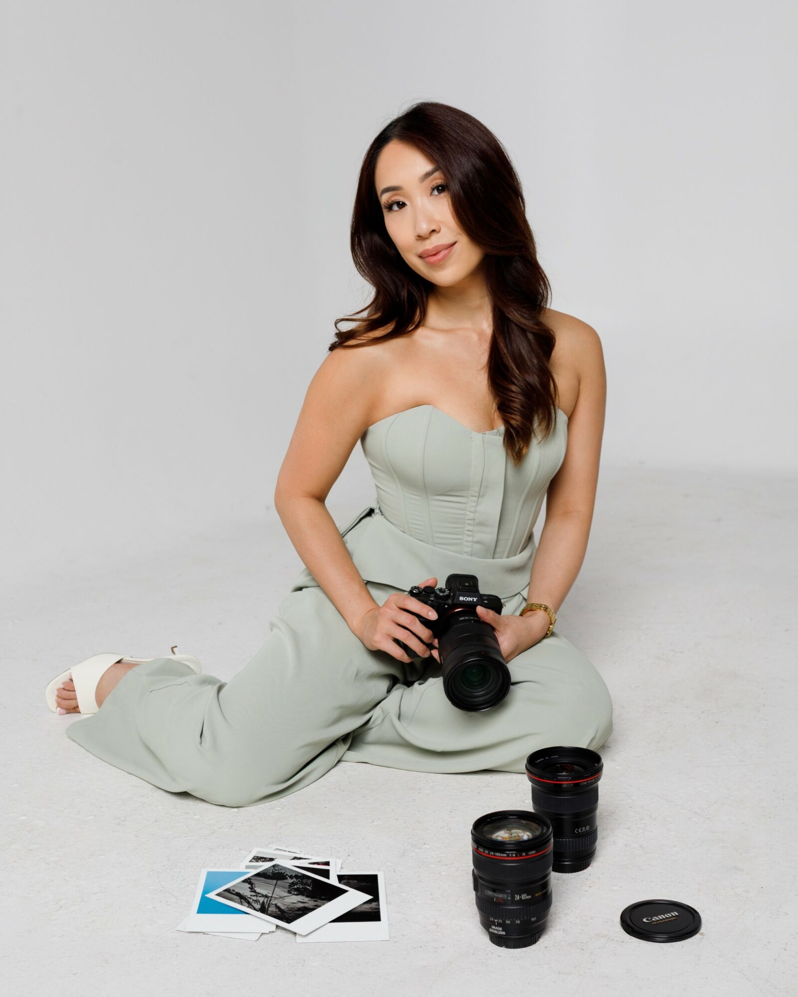 Woman sitting on the floor holding a camera, with camera lenses and photographs nearby, against a plain background.