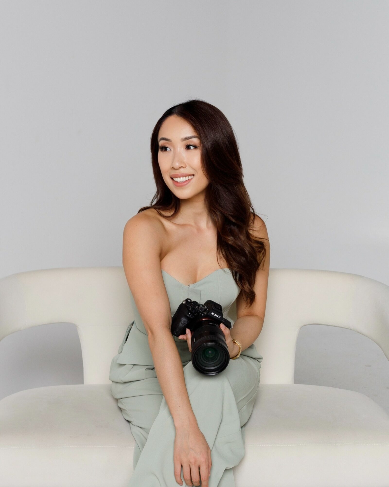 Young woman with long dark hair sitting on a white sofa, holding a camera, smiling, against a plain light gray background.