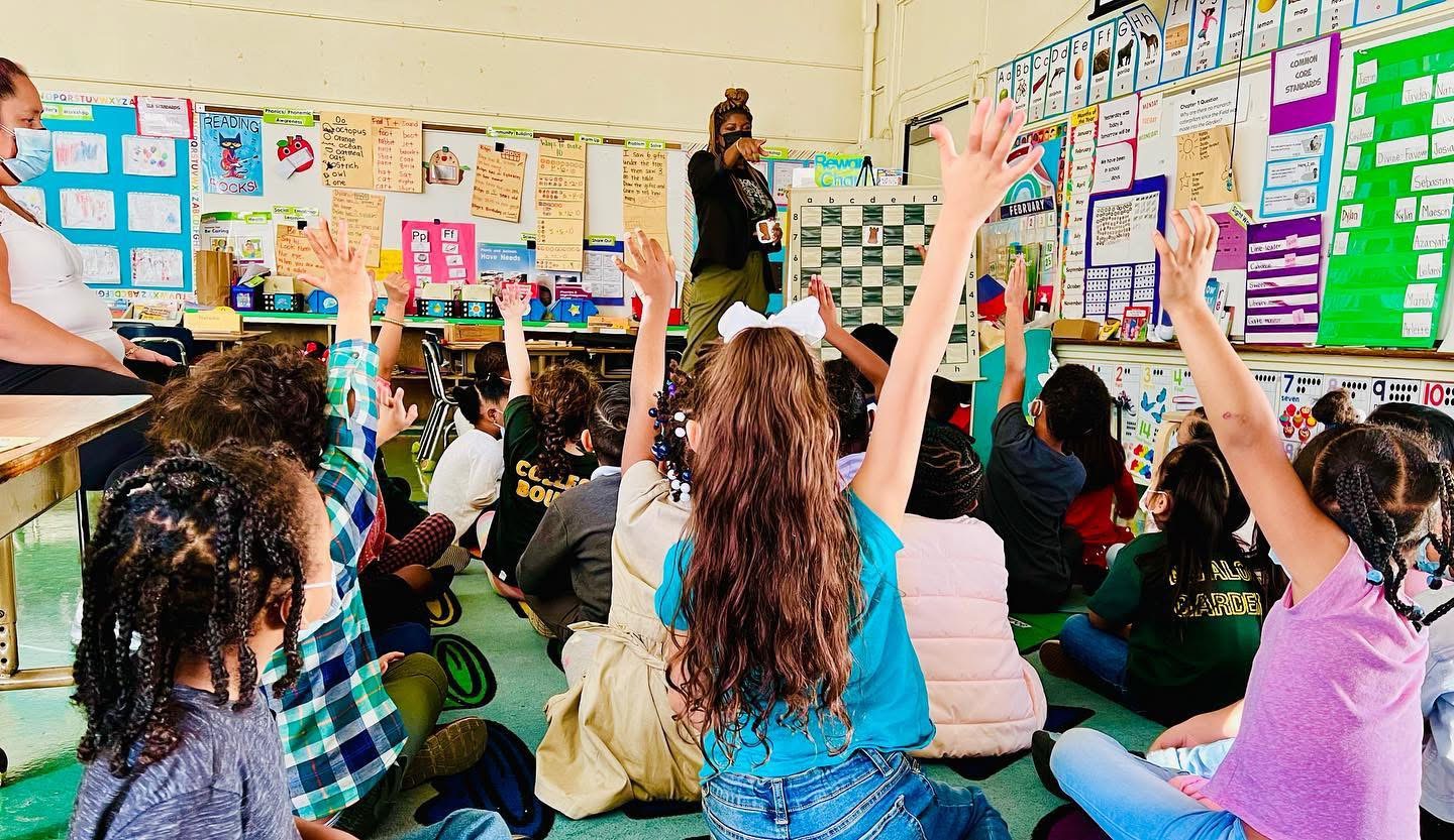 Classroom with students sitting on the floor, raising hands, and a teacher at the front near colorful bulletin boards.