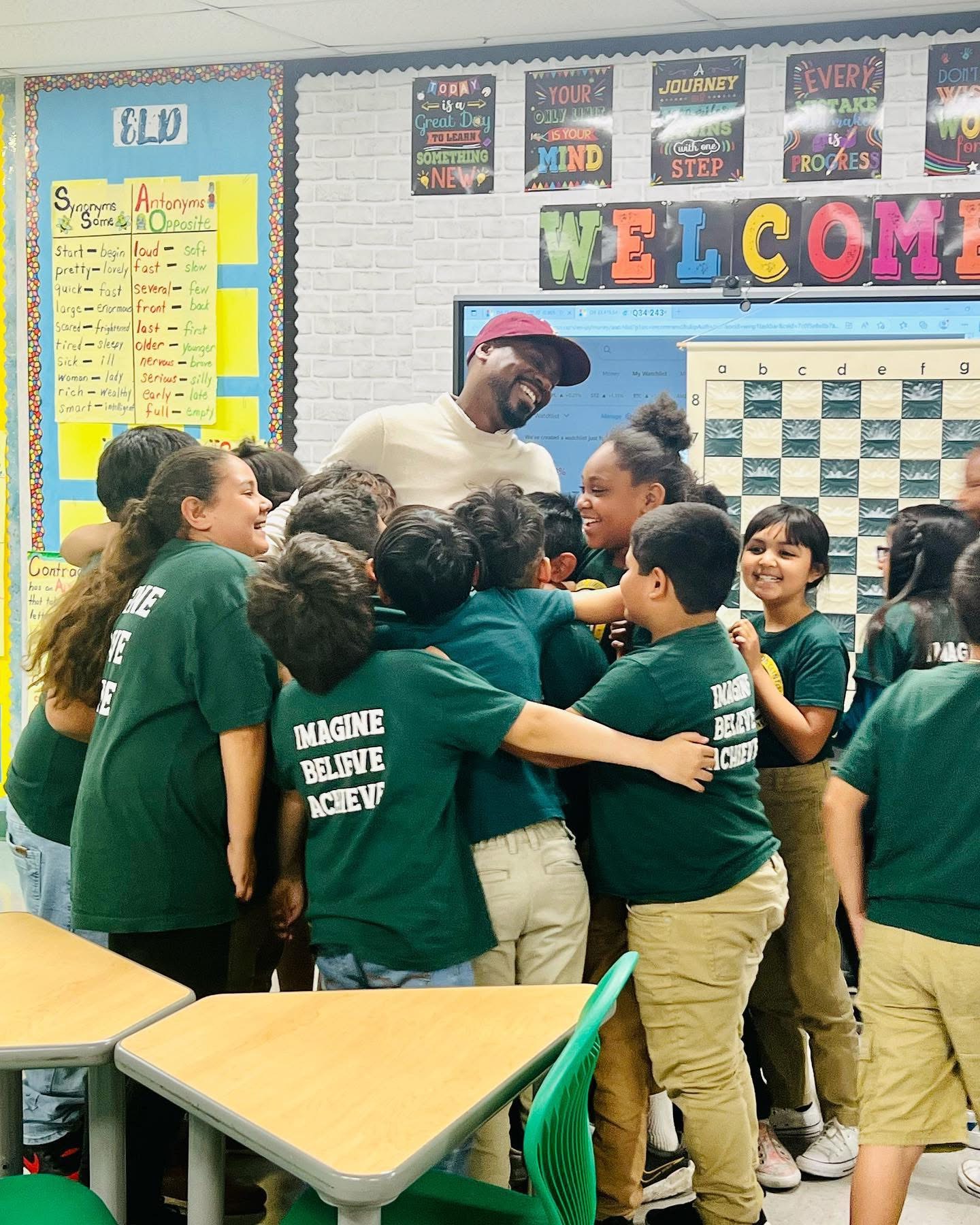 Group of children in green shirts hugging and smiling around an adult in a classroom with colorful decorations and a welcome sign.
