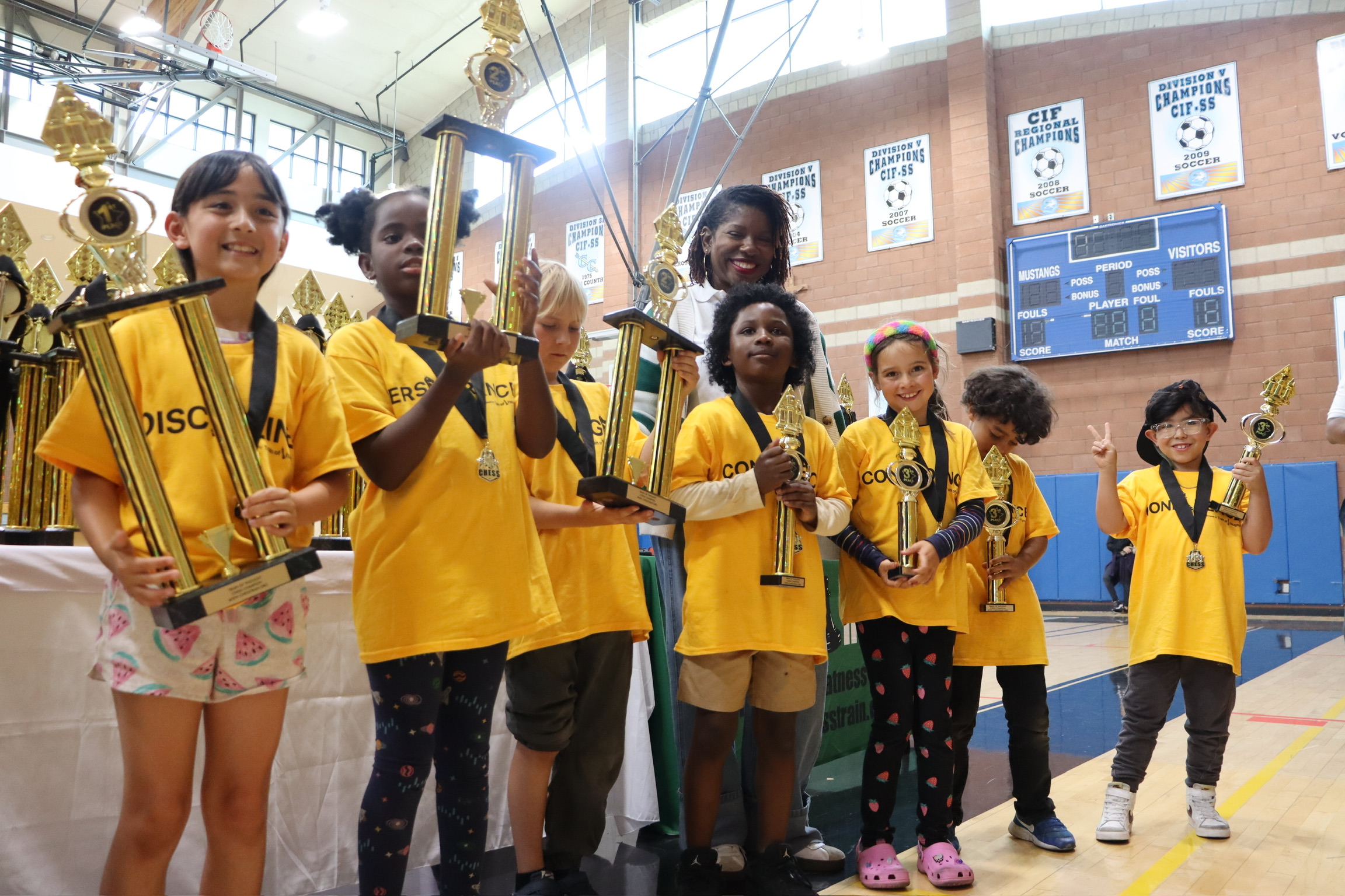 Group of children in yellow shirts holding trophies and medals in a gymnasium, smiling and celebrating.