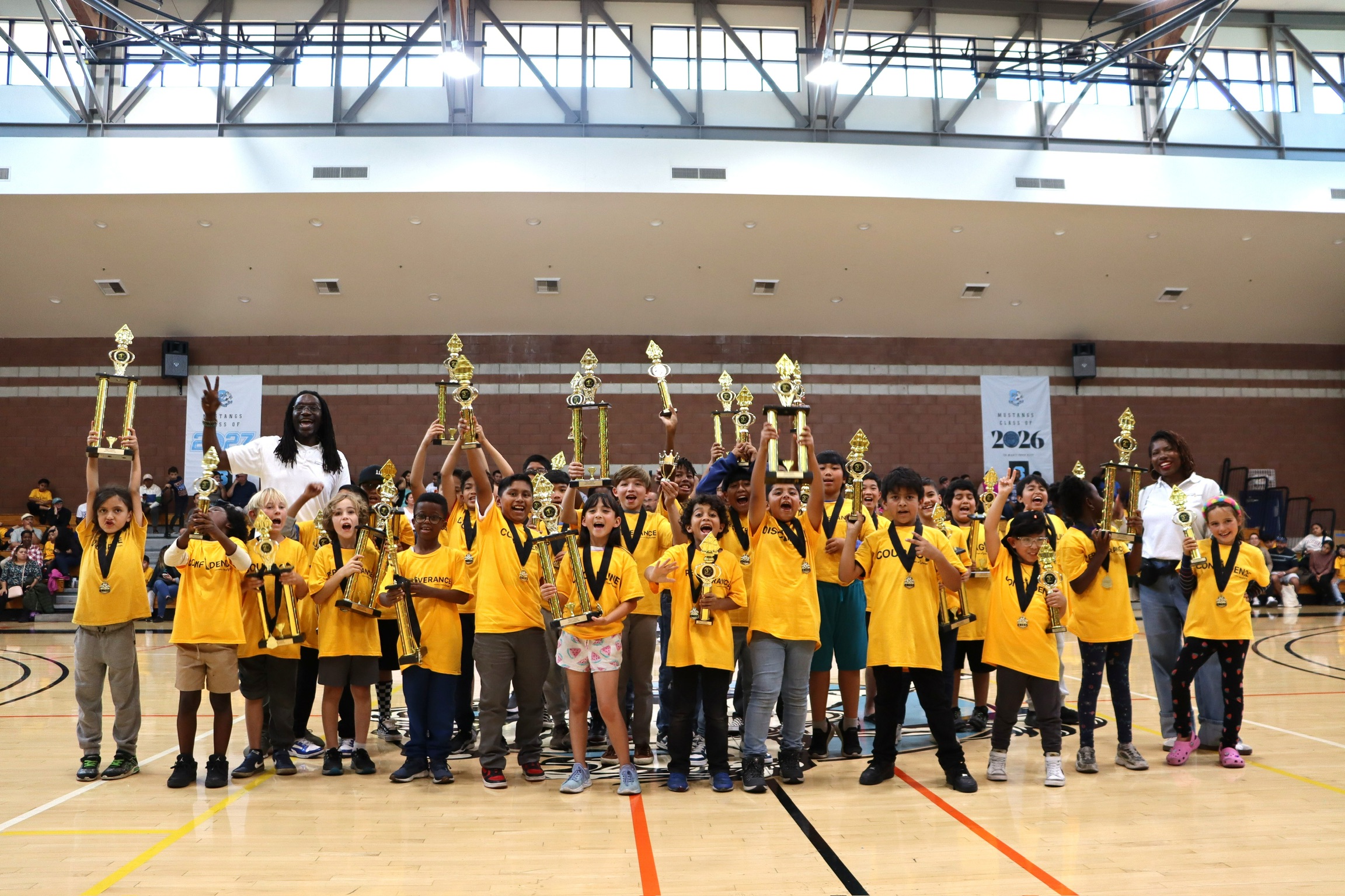 Group of children in yellow shirts holding trophies in a gymnasium, celebrating victory.