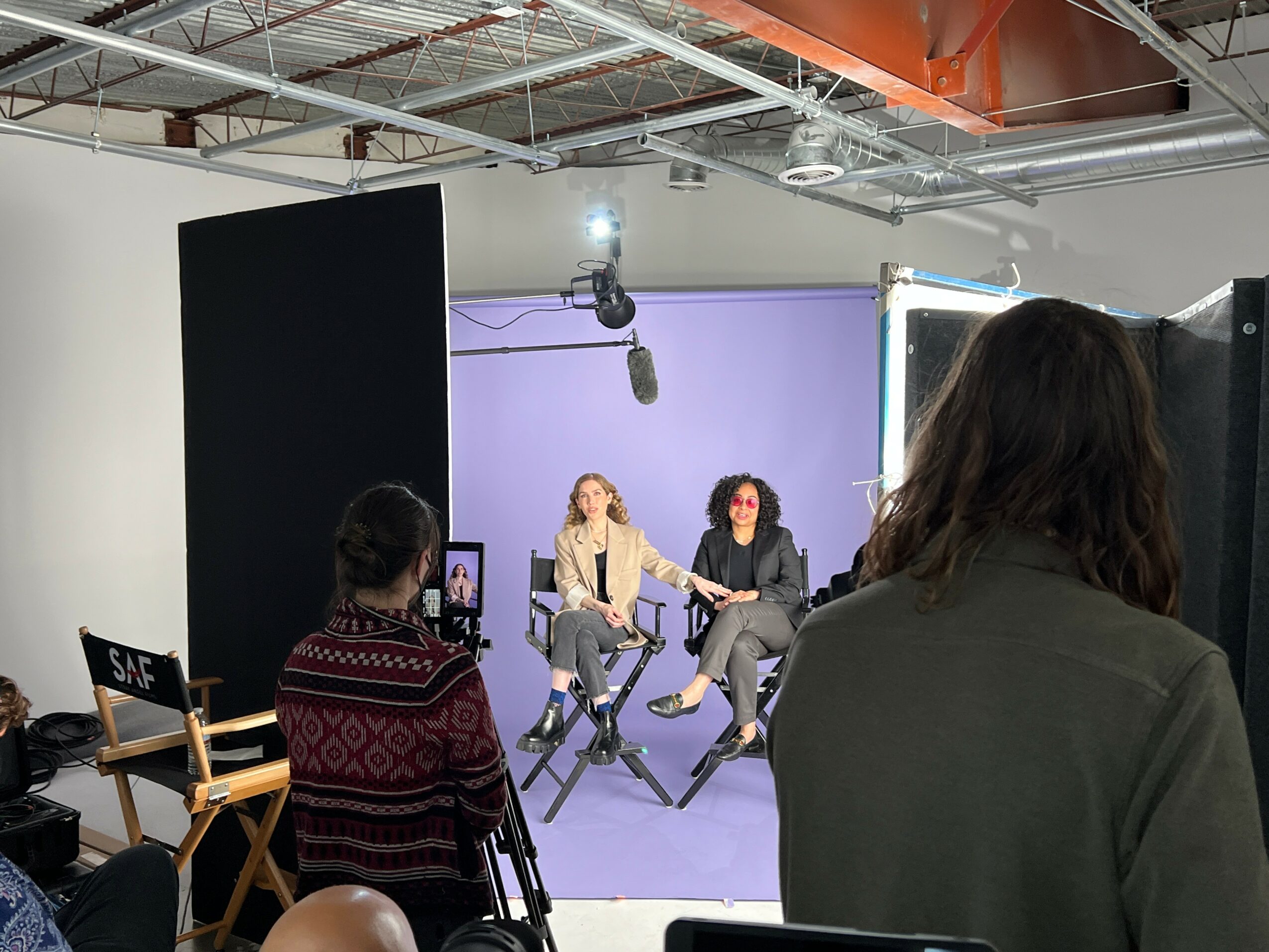 Two women sit on chairs in a studio with purple background, being filmed by a camera and lighting equipment, while two others observe.